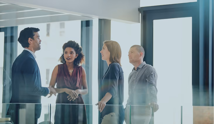 Four business professionals engaged in conversation in a modern office with glass walls and large windows.