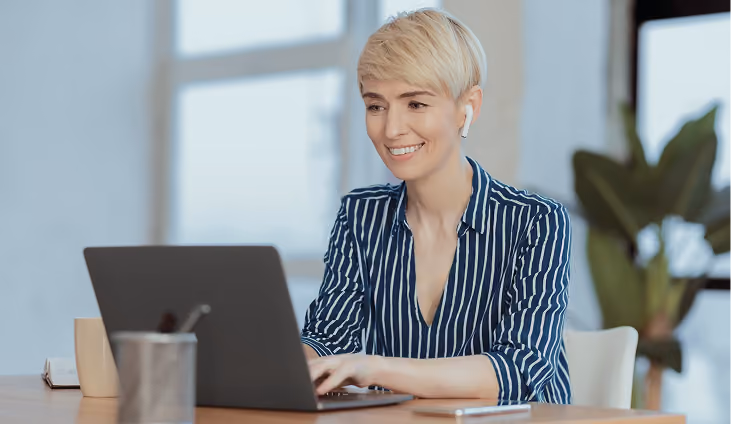 Smiling woman with short blonde hair wearing a blue and white striped shirt, using a laptop at a desk indoors with a blurred plant and window in the background.