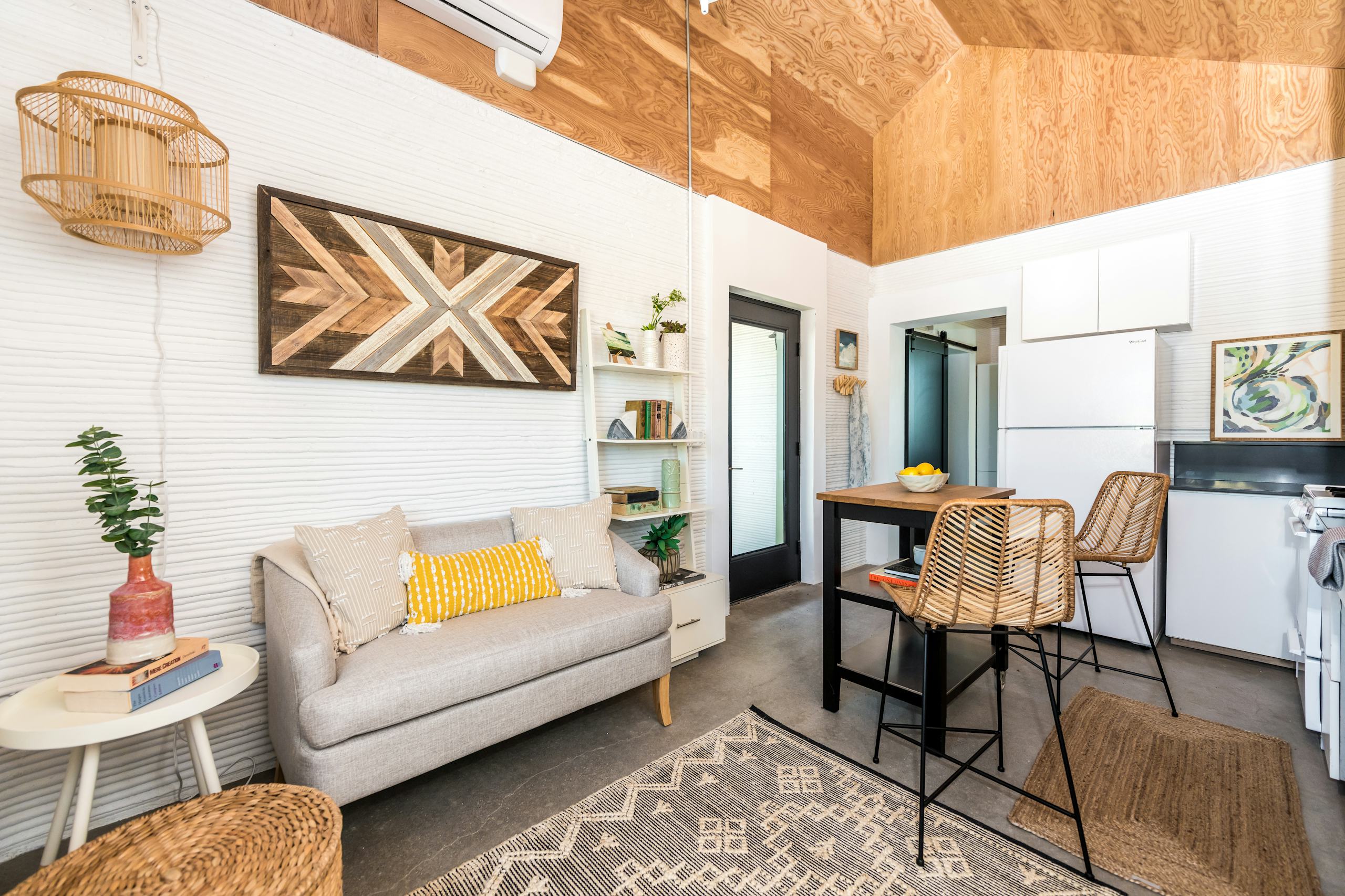 Compact living area with textured 3D-printed white walls, plywood ceiling, kitchenette and rattan bar stools