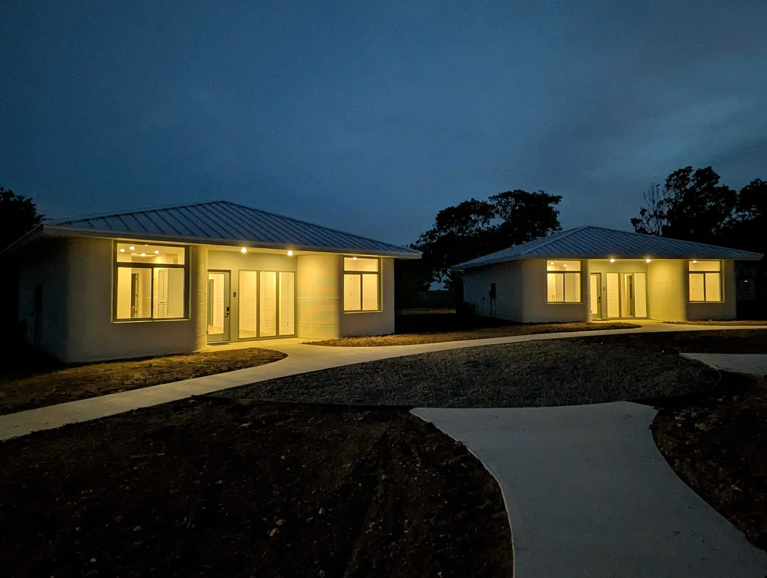 Night view of two single‑story modern homes with illuminated entries and winding concrete paths in a residential community