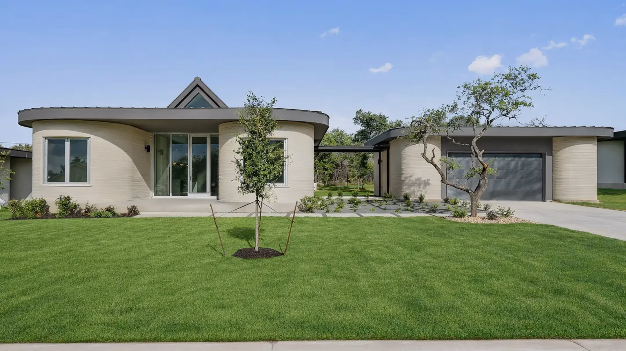 Modern 3D-printed home with curved walls, central glass entrance, garage and front lawn