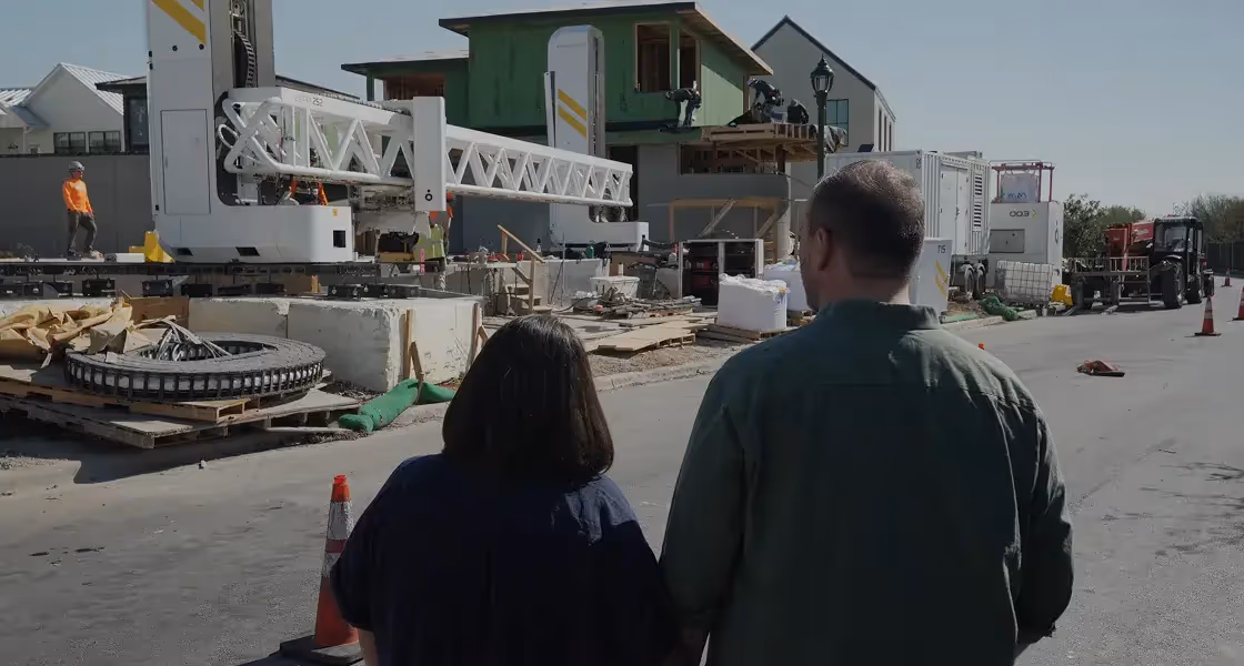 Two people from behind near a construction site with a large white 3D printer and partially built house