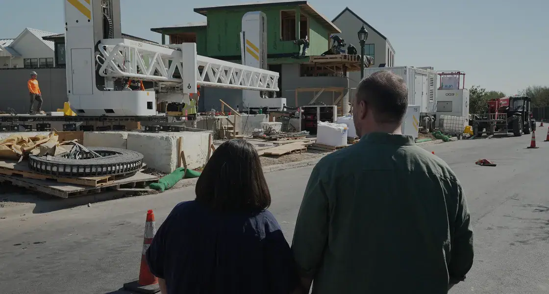 Two people from behind near a construction site with a large white 3D printer and partially built house
