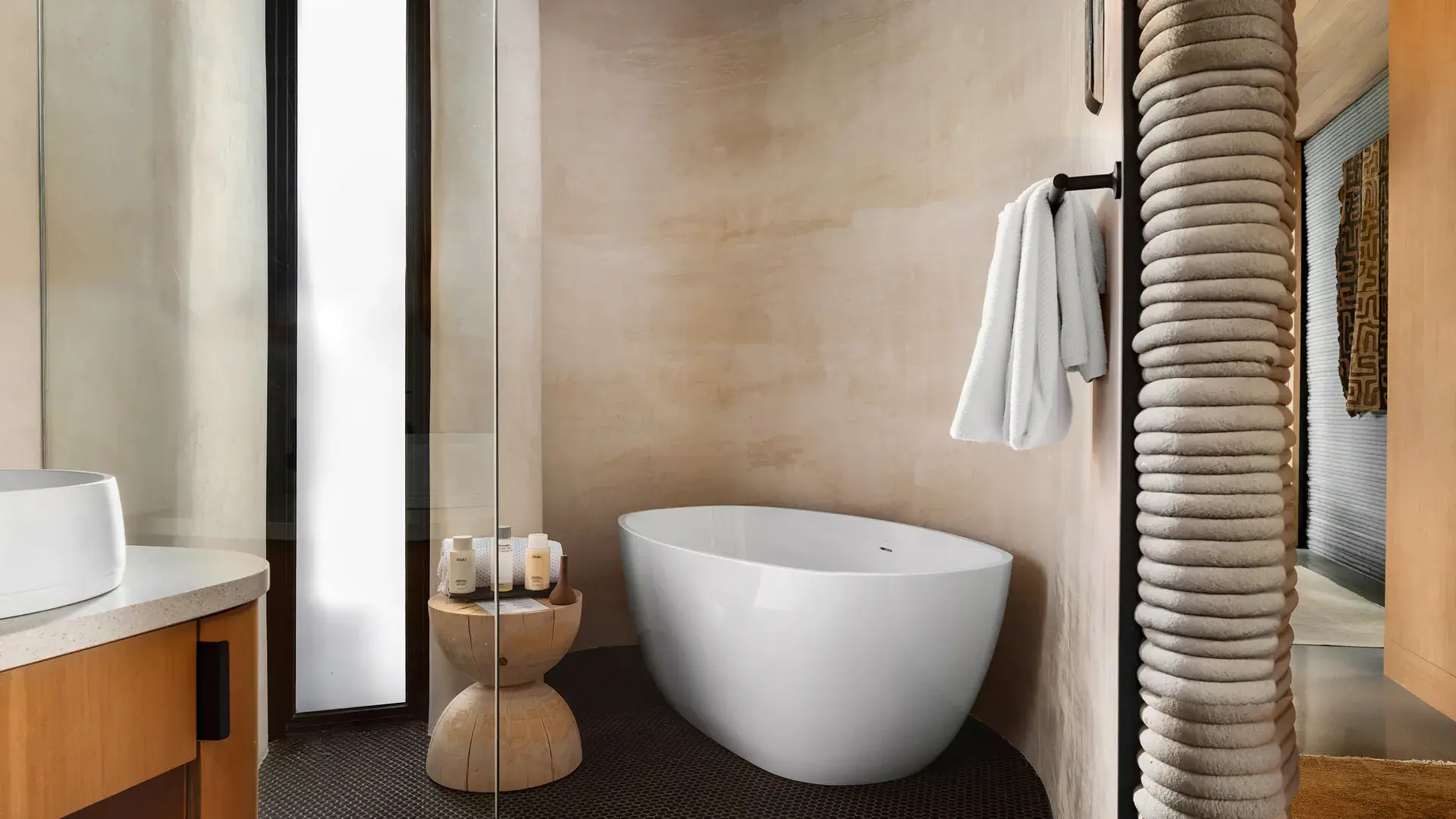White freestanding bathtub in a minimalist bathroom with wooden stool, toiletries, and towel