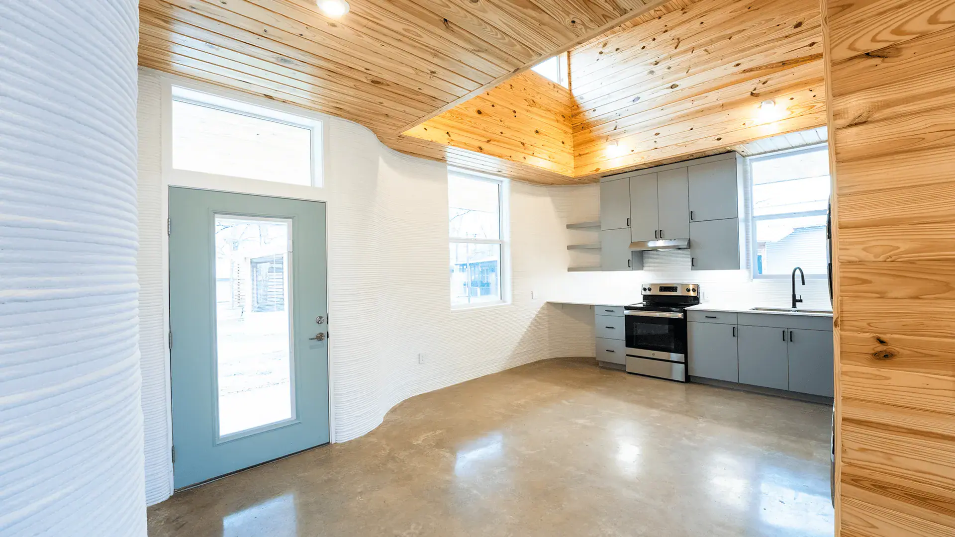 Open kitchen with ribbed white 3D-printed walls, wood ceiling, gray cabinets, and blue door
