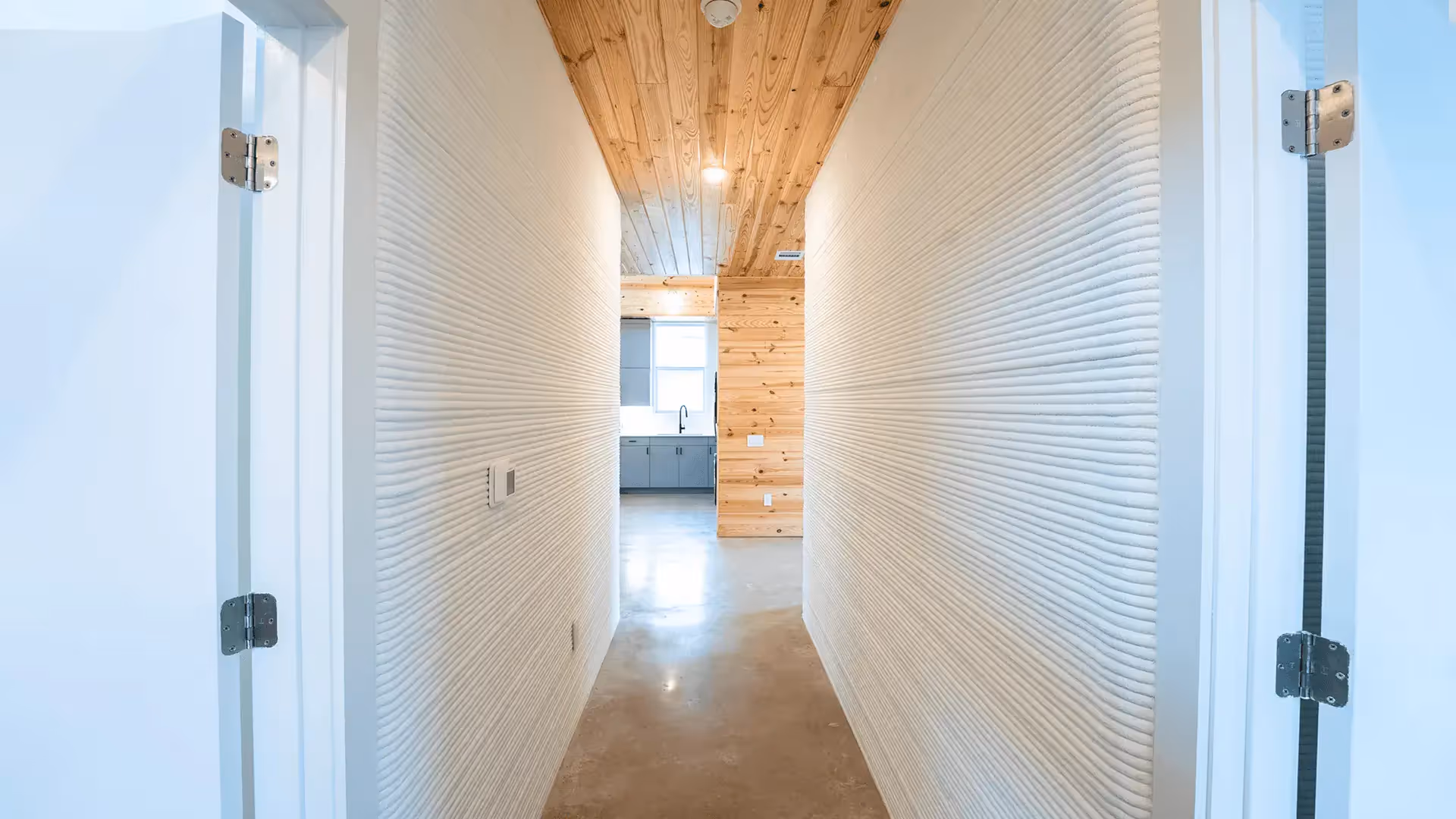Narrow hallway with ribbed white 3D-printed walls, wood ceiling and concrete floor, view into kitchen