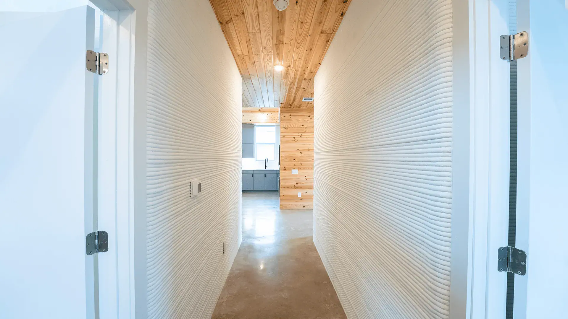 Narrow hallway with ribbed white 3D-printed walls, wood ceiling and concrete floor, view into kitchen