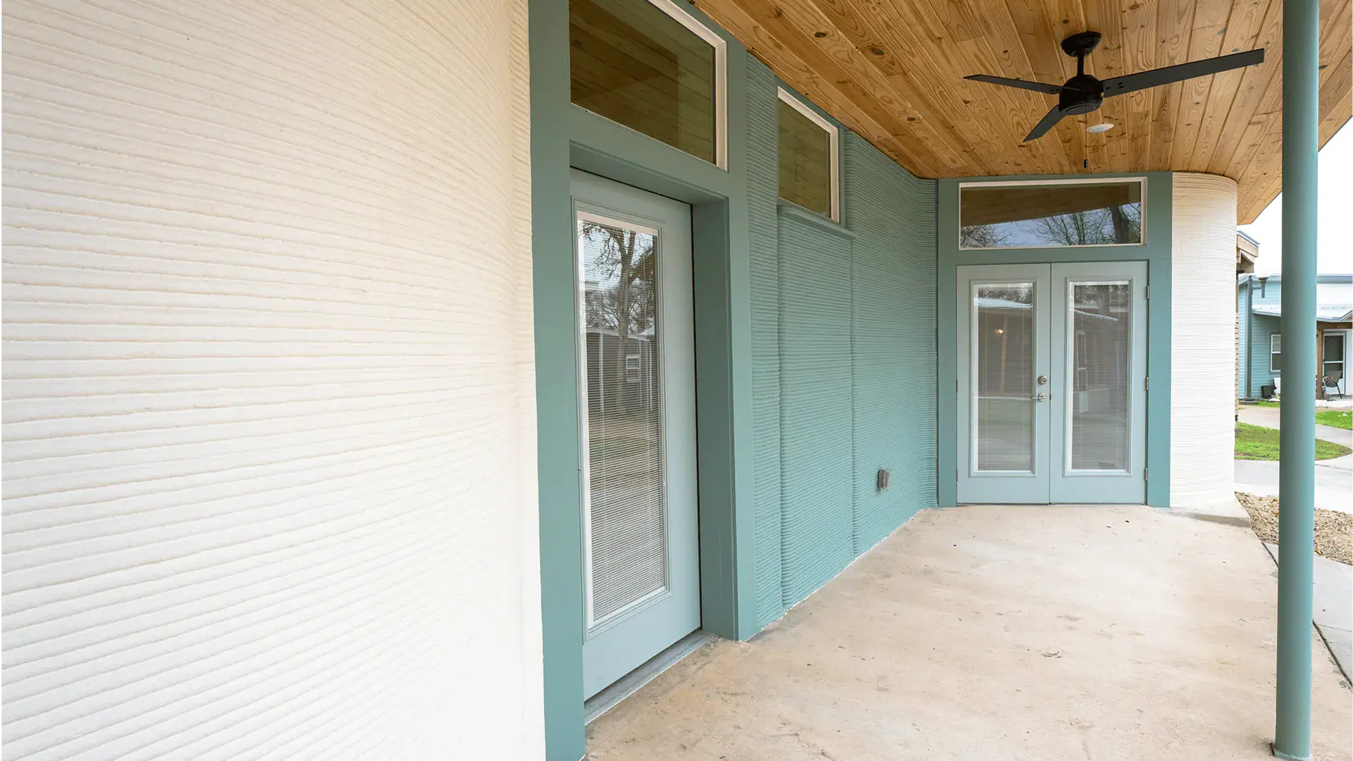 Covered porch with teal doors, ribbed 3D-printed walls, wood-paneled ceiling and black fan