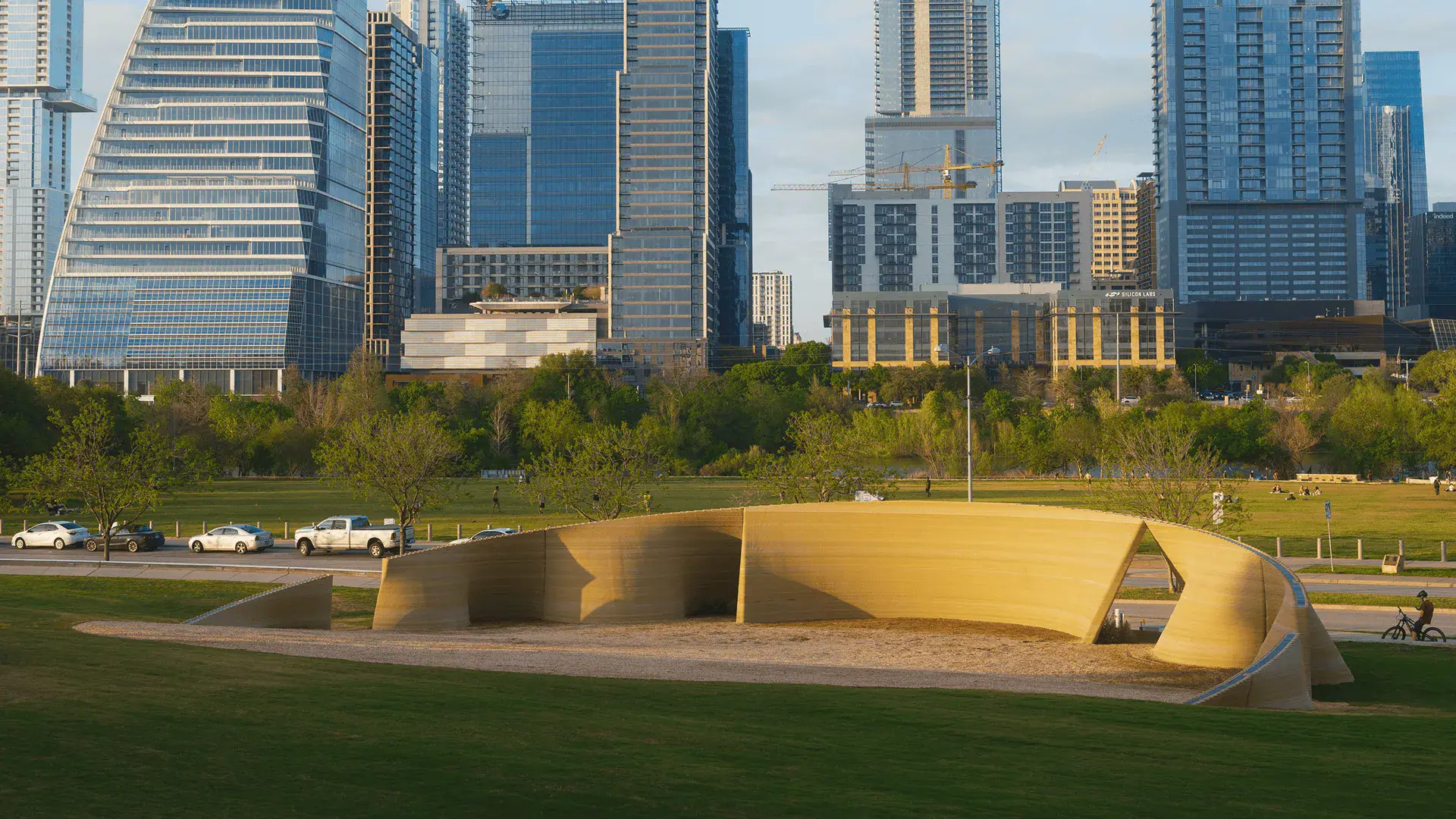 Curved 3D-printed concrete pavilion in a park with downtown skyline