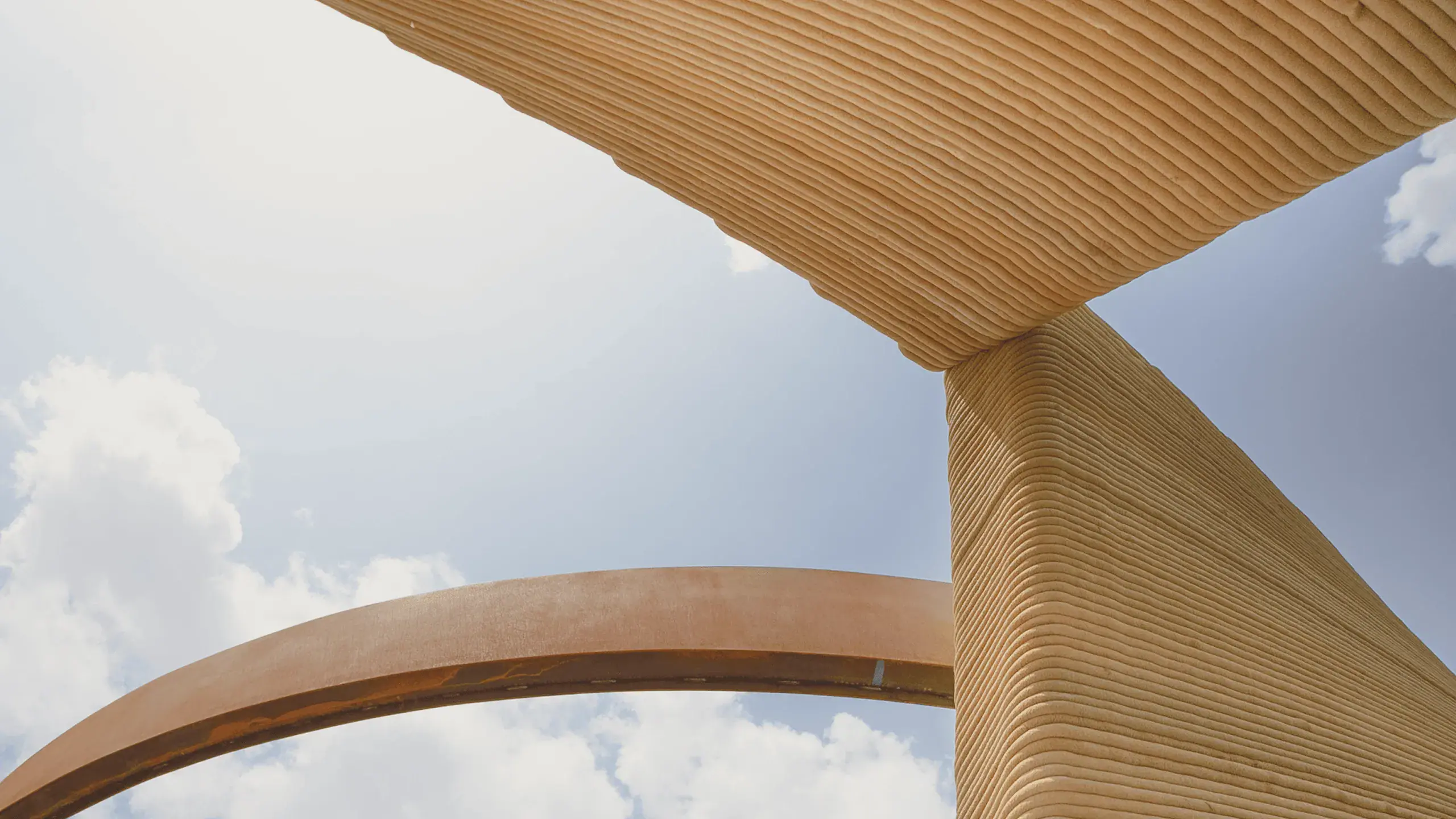 Layered 3D-printed concrete wall and slab meeting above a rusted metal arch against blue sky