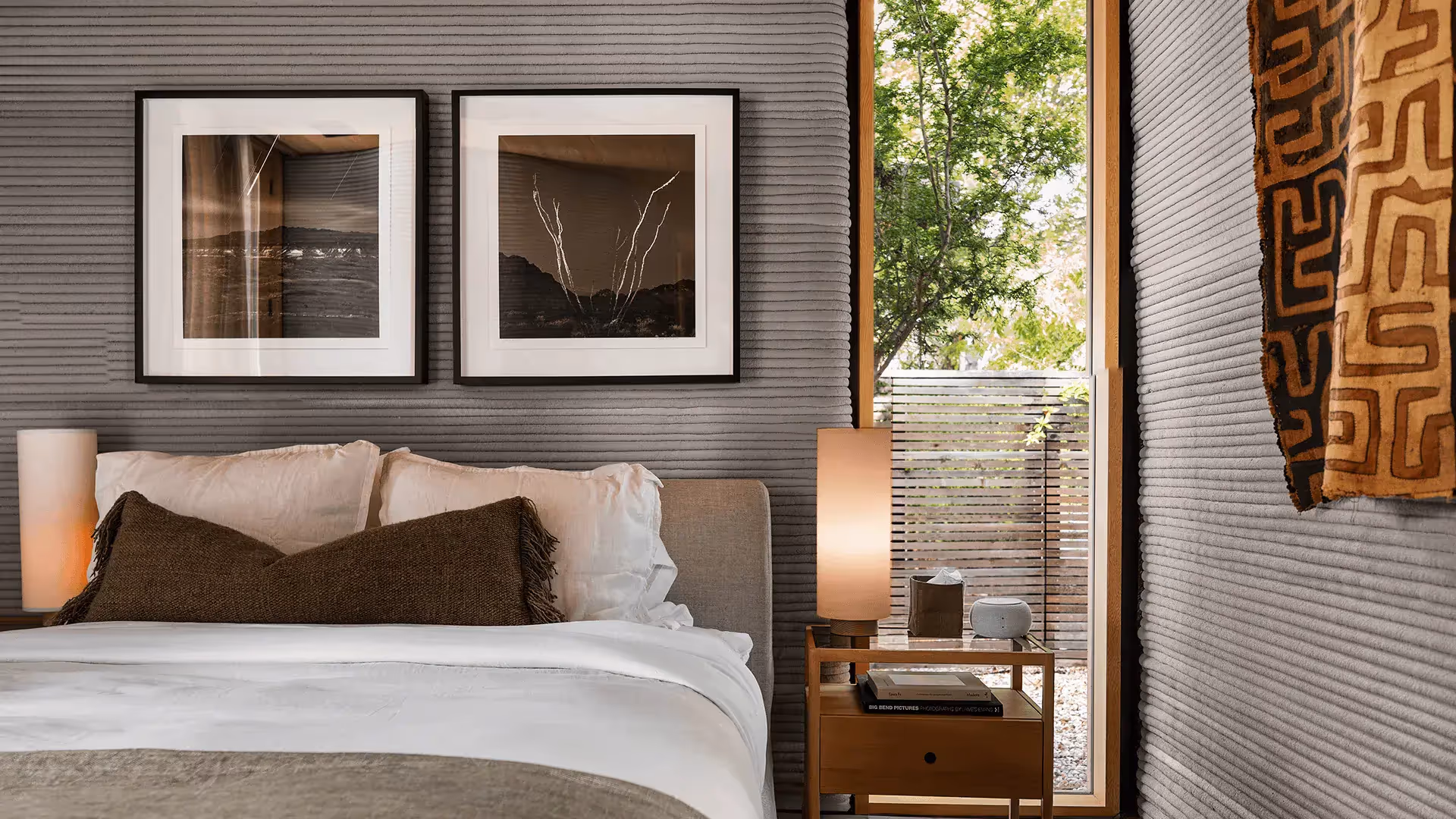 Bedroom with white bedding, brown lumbar pillow, ribbed gray walls, framed art, nightstand lamp, and window with trees