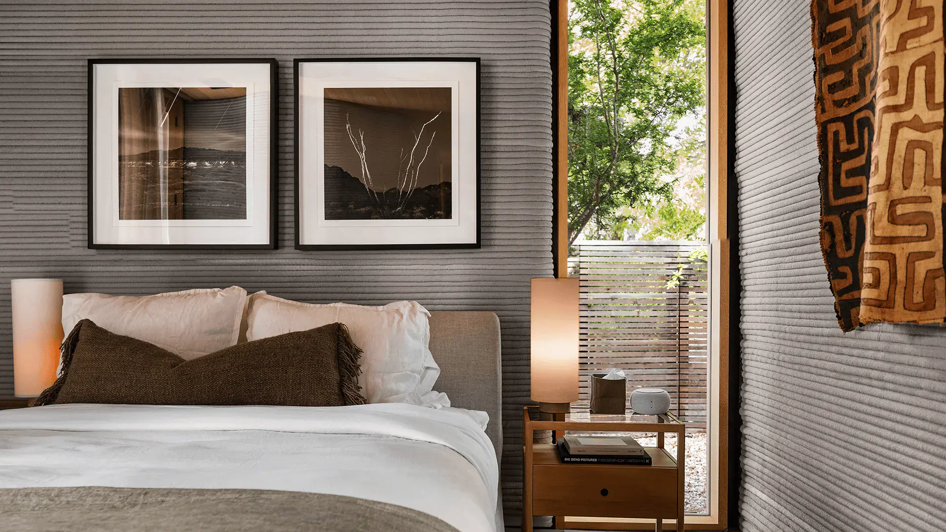 Bedroom with white bedding, brown lumbar pillow, ribbed gray walls, framed art, nightstand lamp, and window with trees
