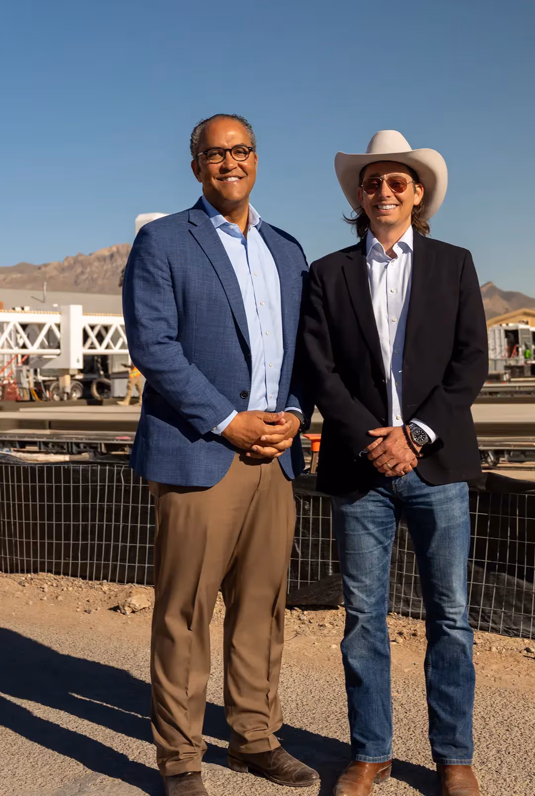 Two company leaders at a 3D‑printed housing construction site with robotic printing equipment and desert mountains behind