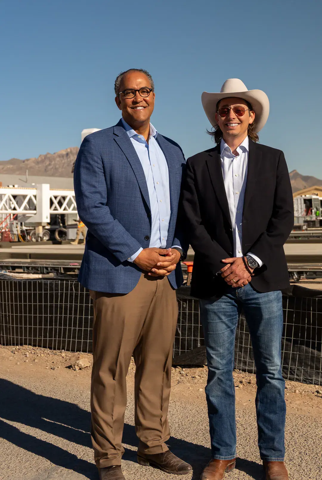 Two company leaders at a 3D‑printed housing construction site with robotic printing equipment and desert mountains behind