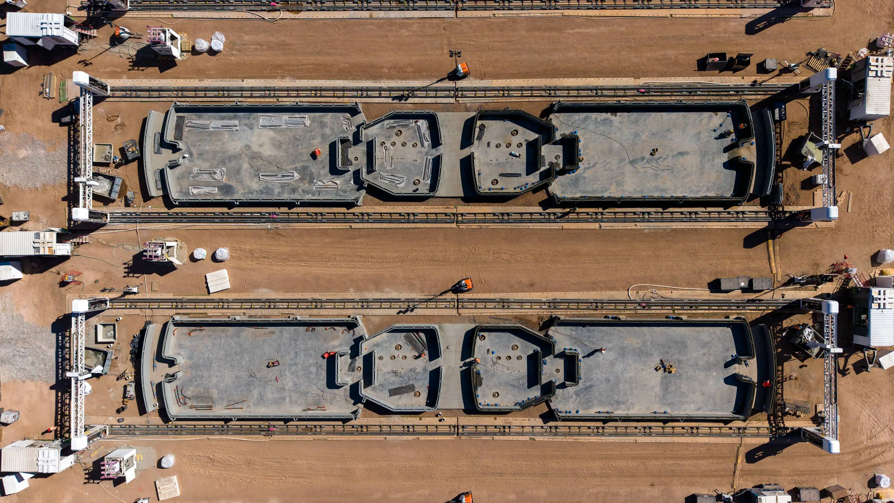 Aerial view of dual gantry robotic 3D printers printing modular concrete home foundations on a sandy construction site