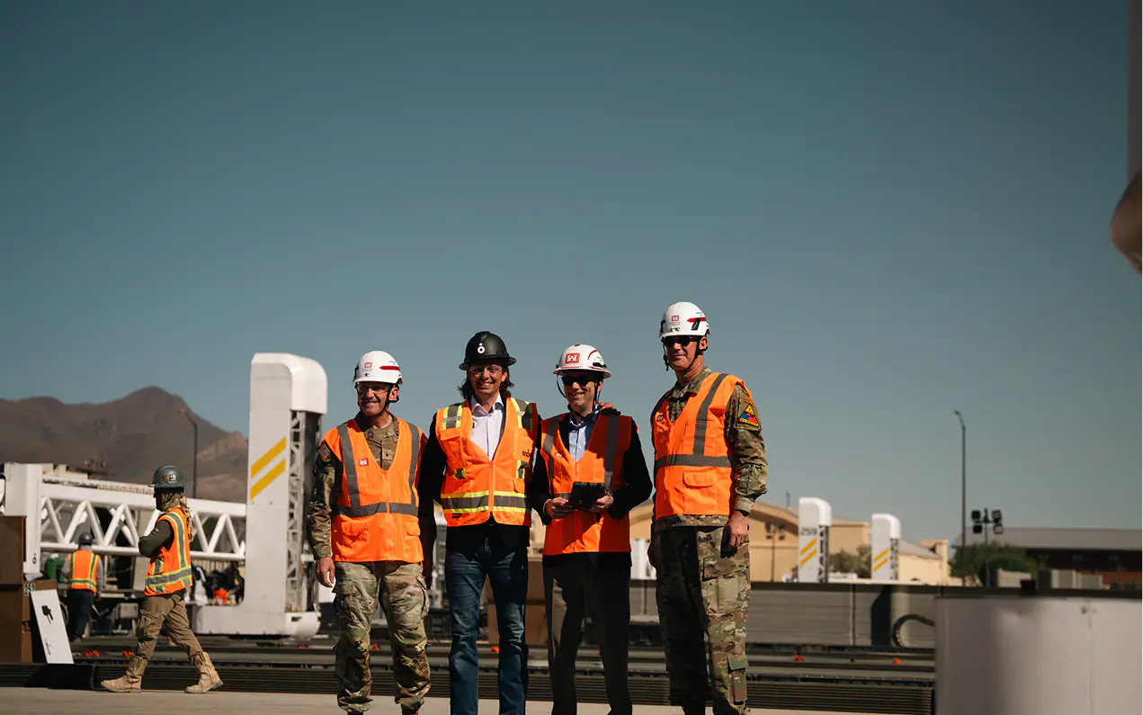 Four people in hard hats and orange safety vests at a construction site with robotic printing equipment behind them