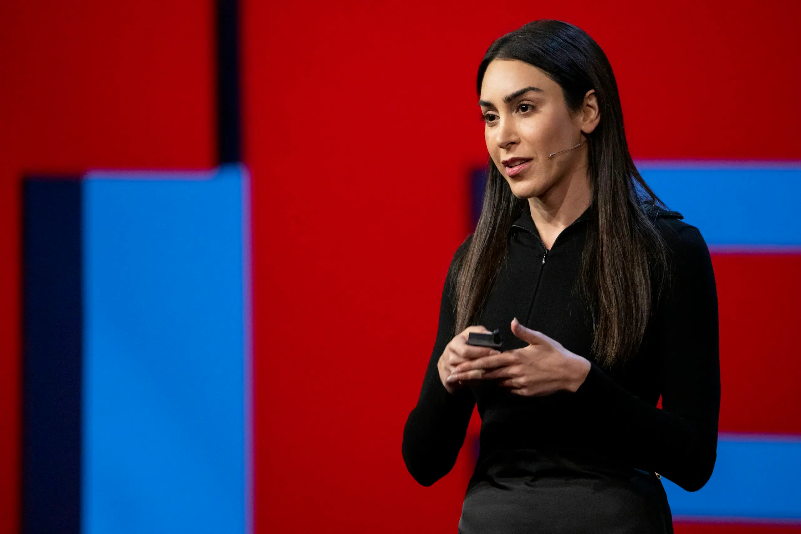 Woman presenter with headset microphone holding a clicker on stage against a red and blue backdrop