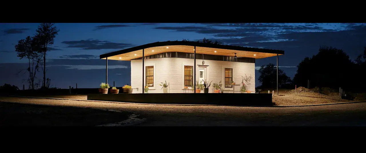 3D-printed single-story home prototype with cantilevered roof and lit porch at dusk, showcasing construction technology