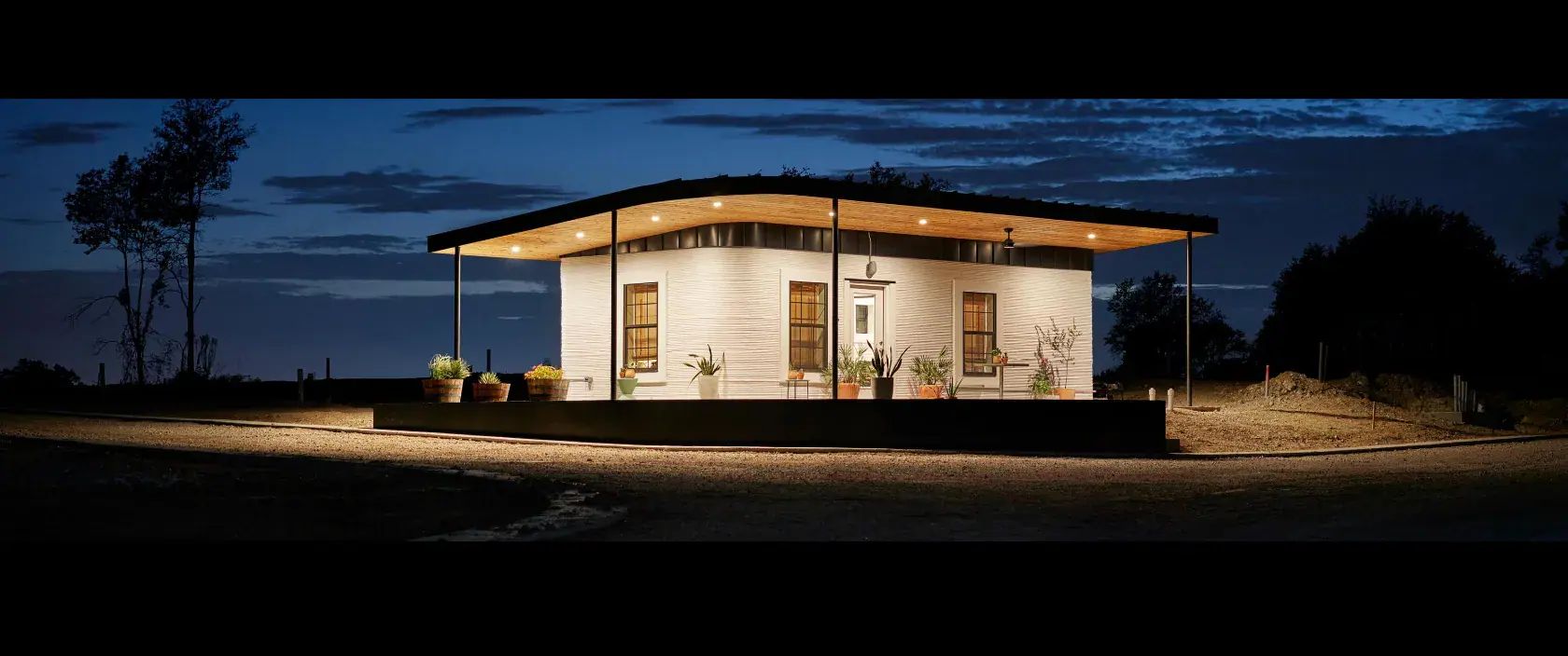 3D-printed single-story home prototype with cantilevered roof and lit porch at dusk, showcasing construction technology