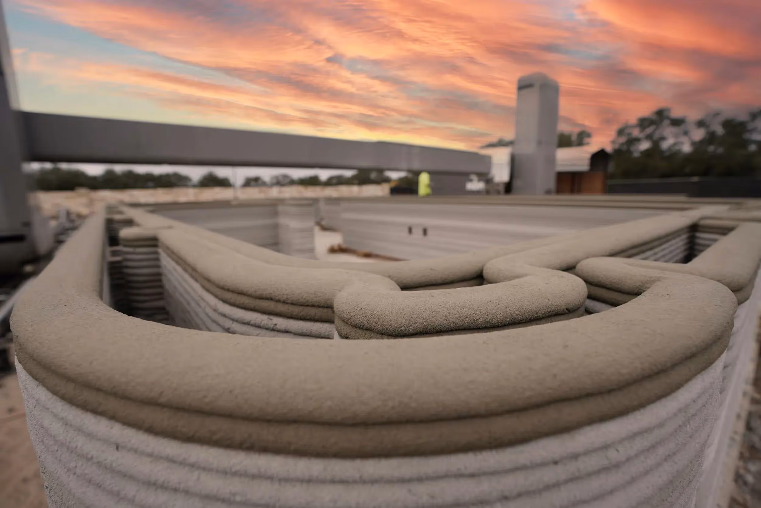 Layered 3D-printed concrete wall on a job site at sunset, illustrating robotic extrusion construction technology