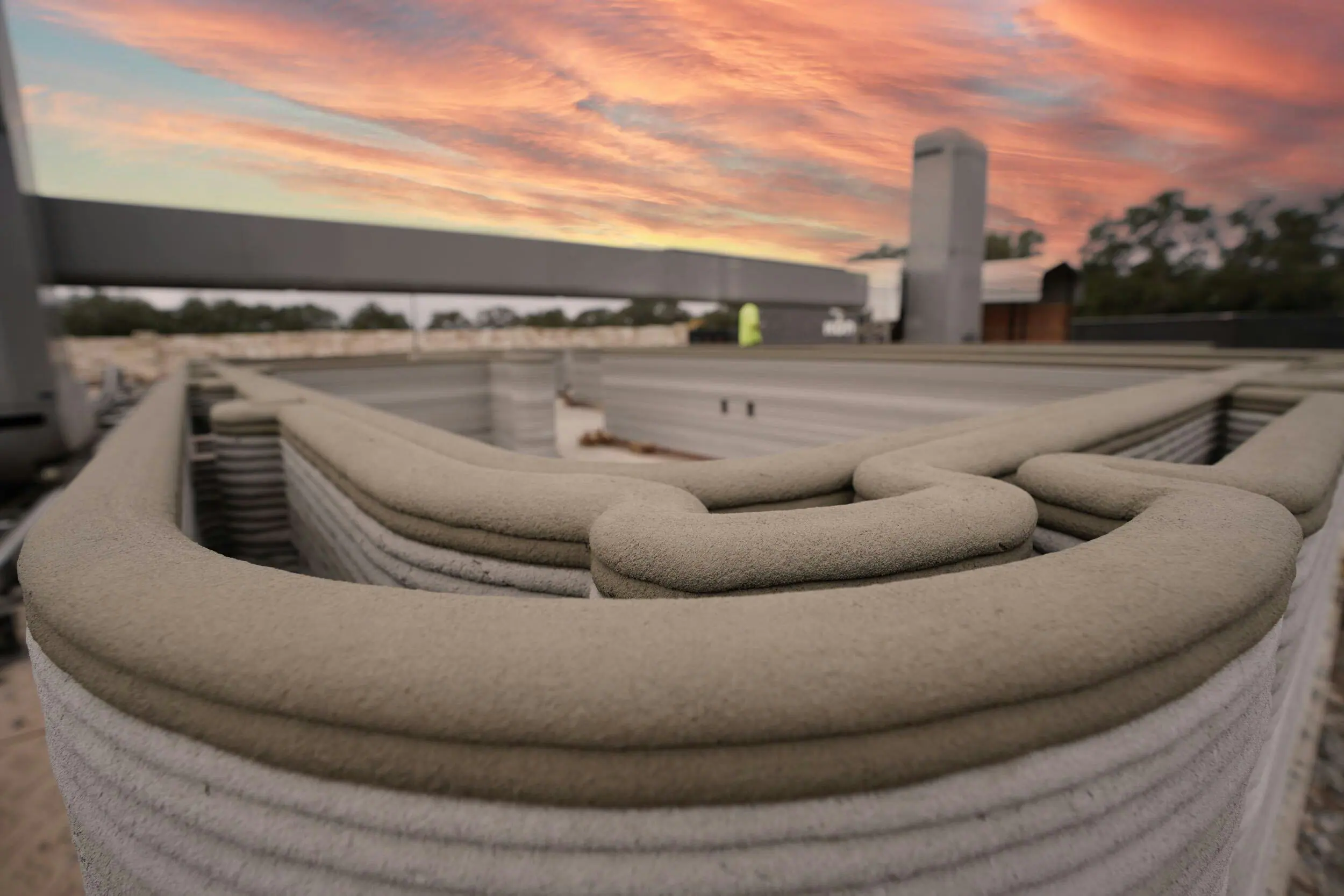 Layered 3D-printed concrete wall on a job site at sunset, illustrating robotic extrusion construction technology
