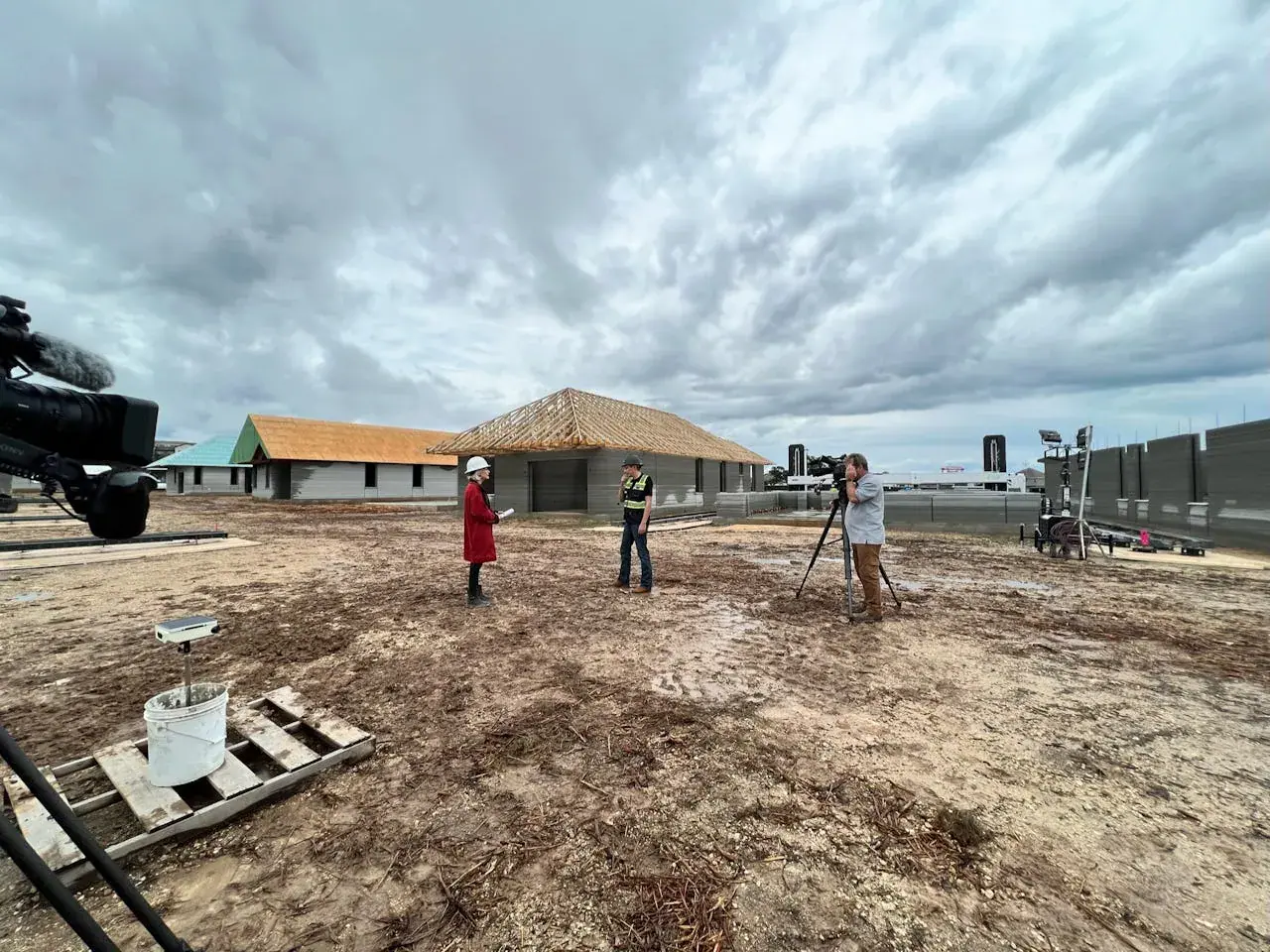 News crew interviewing a manager at a construction tech site with partially built homes and roof framing