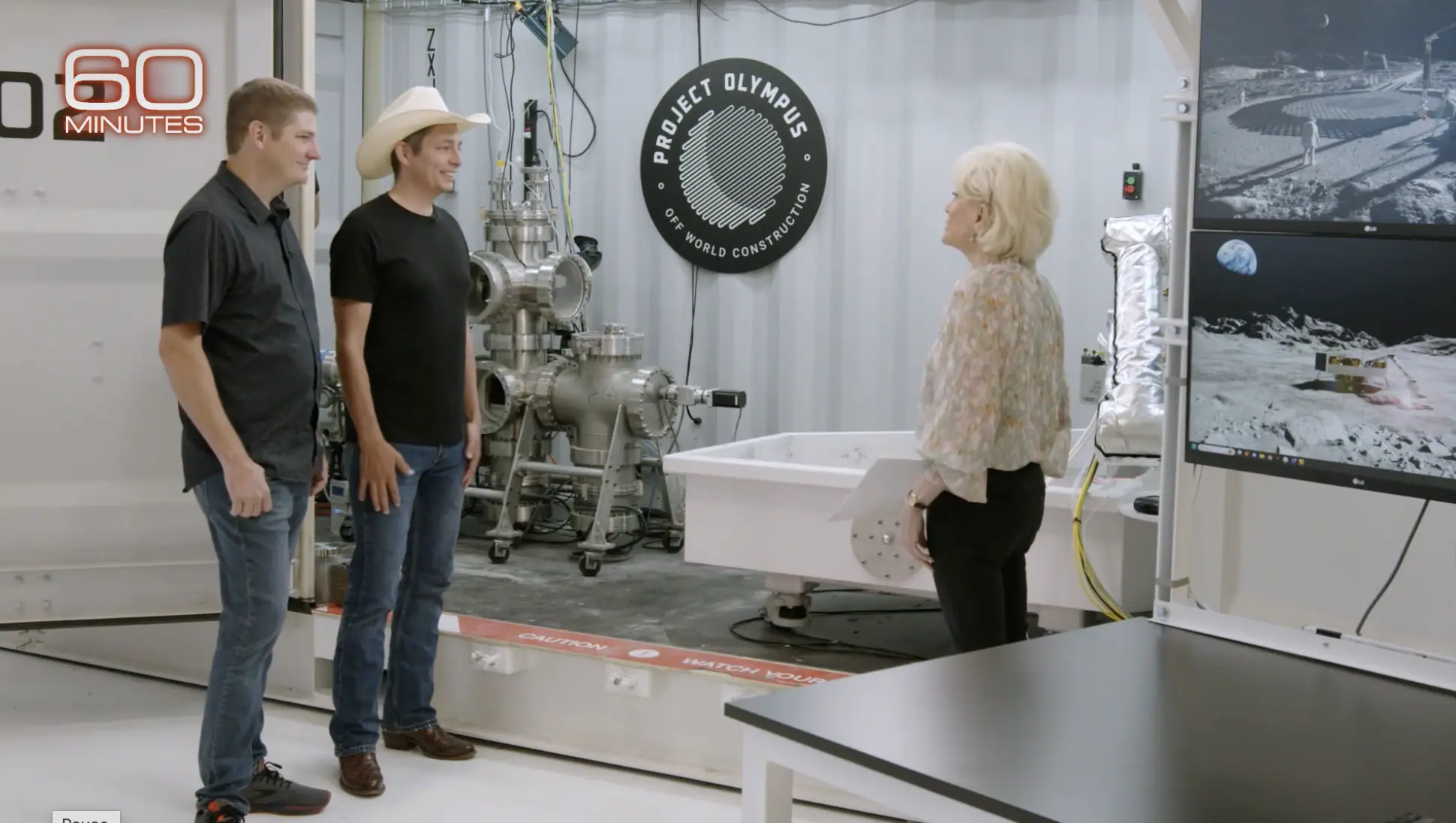 Two men and a woman stand in a construction robotics lab beside Project Olympus sign and lunar display during TV segment