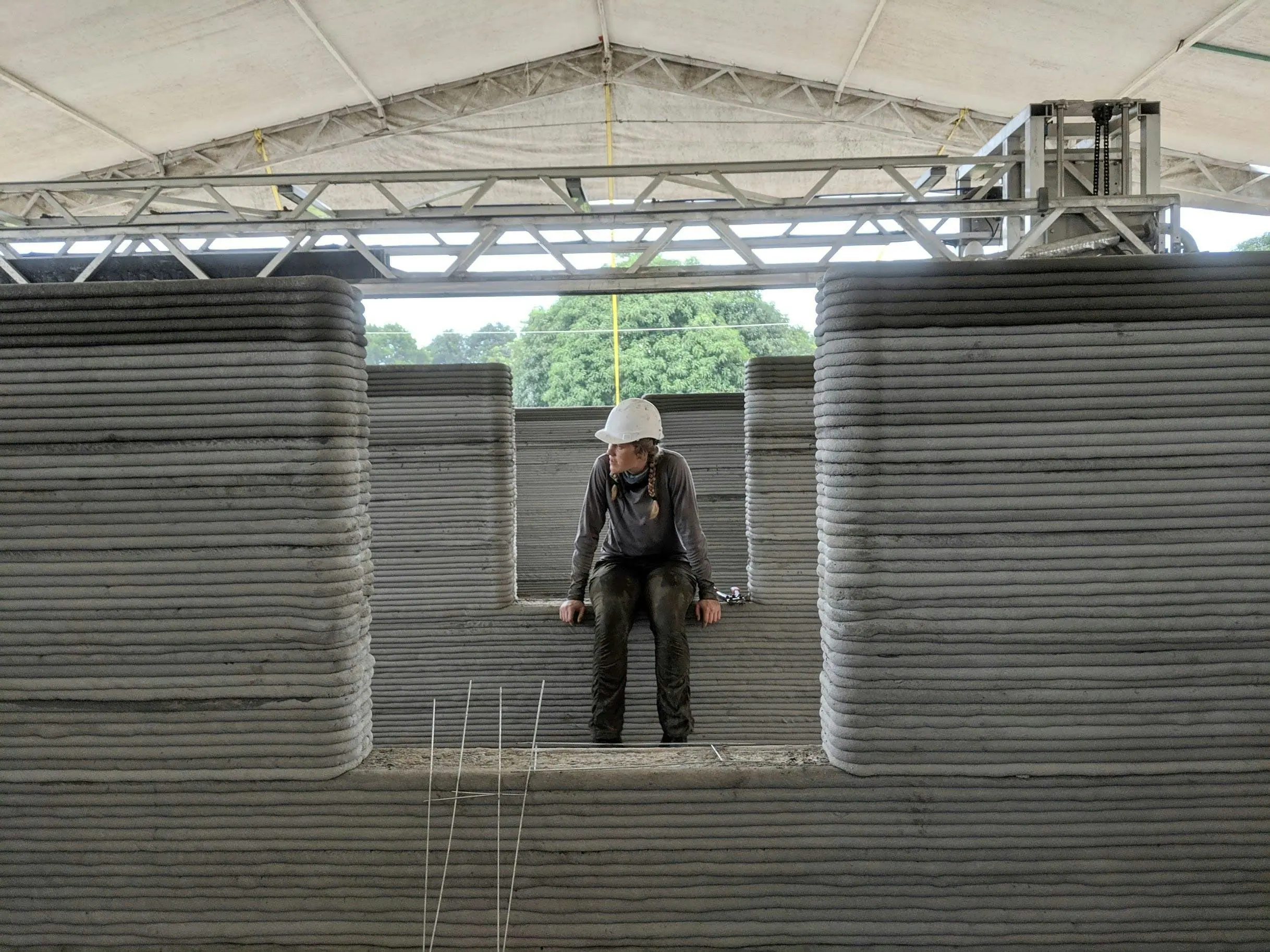 Construction worker in hard hat seated on layered 3D-printed concrete wall sections with overhead truss and exposed rebar