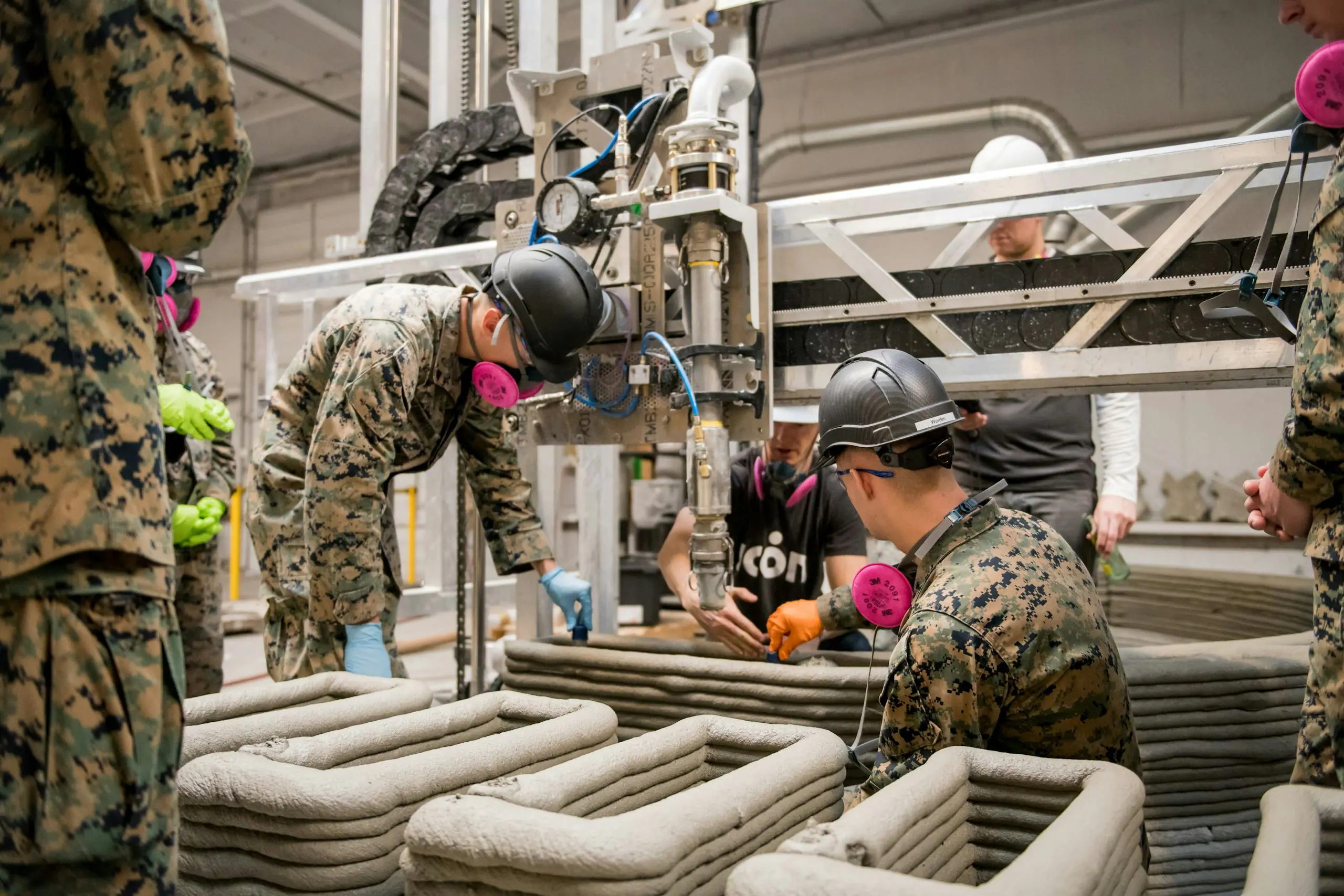 Uniformed crew wearing helmets and respirators operating a robotic 3D concrete printer layering walls