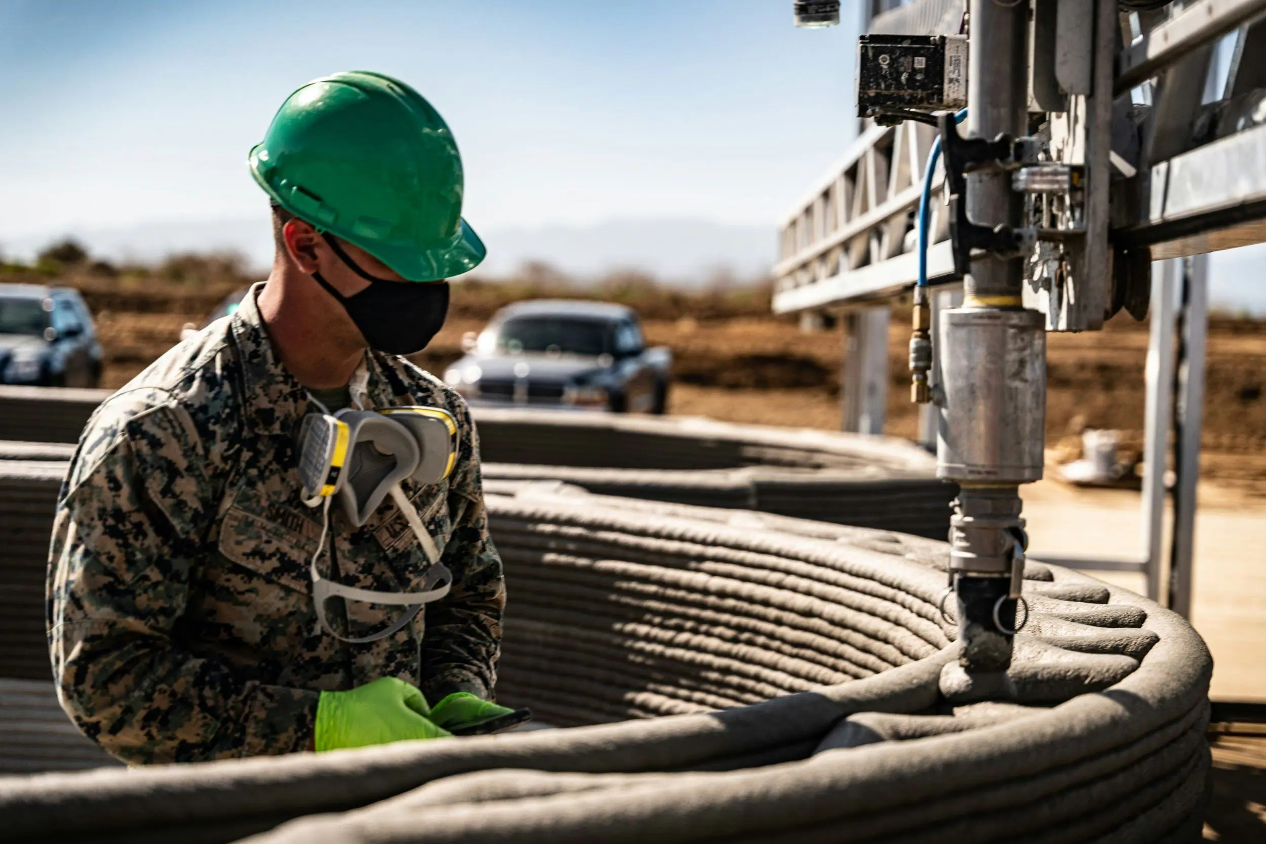 Technician with green hard hat and respirator monitoring robotic nozzle extruding layered concrete for a 3D‑printed wall