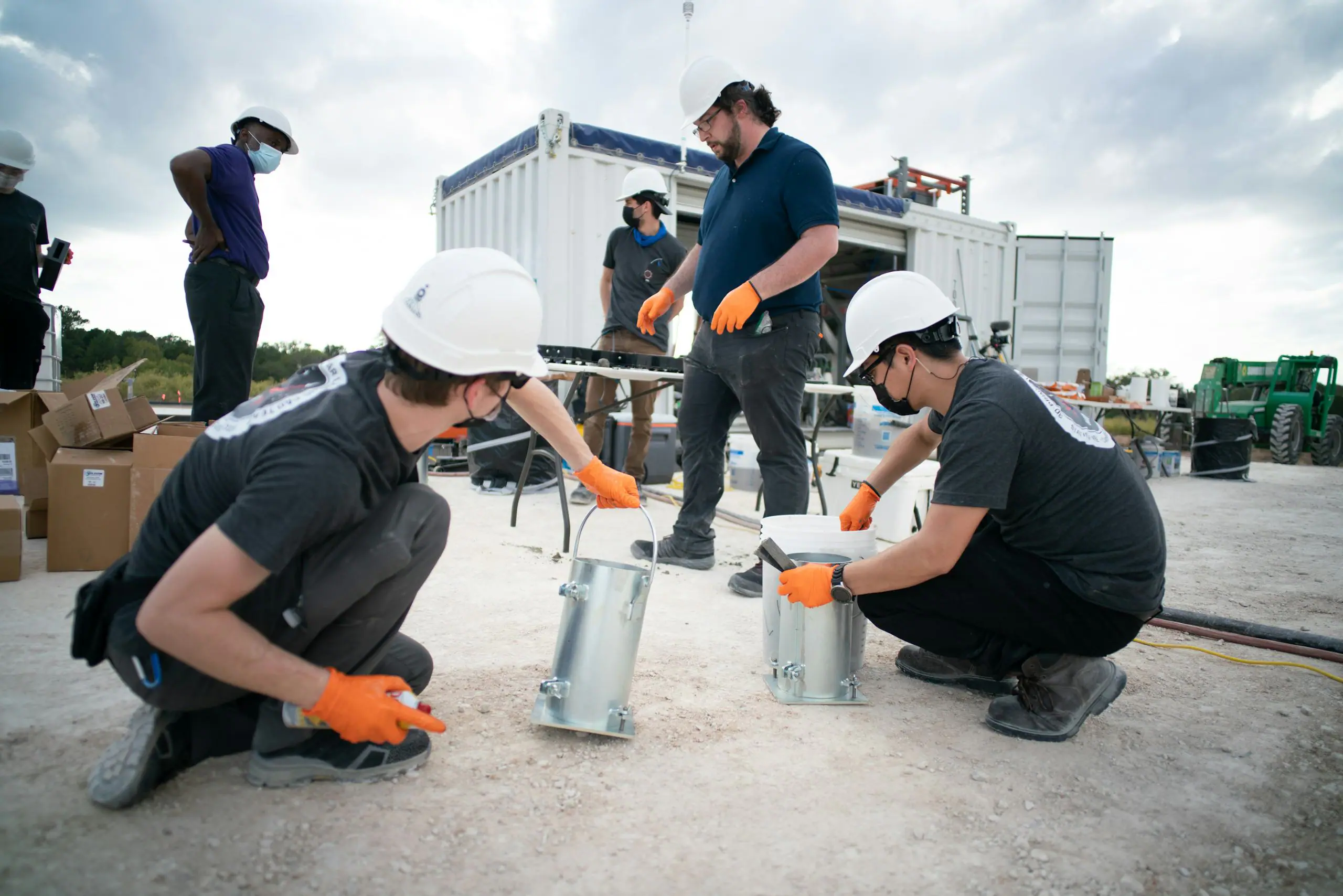 Team in hard hats and orange gloves installing metal footing anchors on site during foundation prep for 3D-printed housing