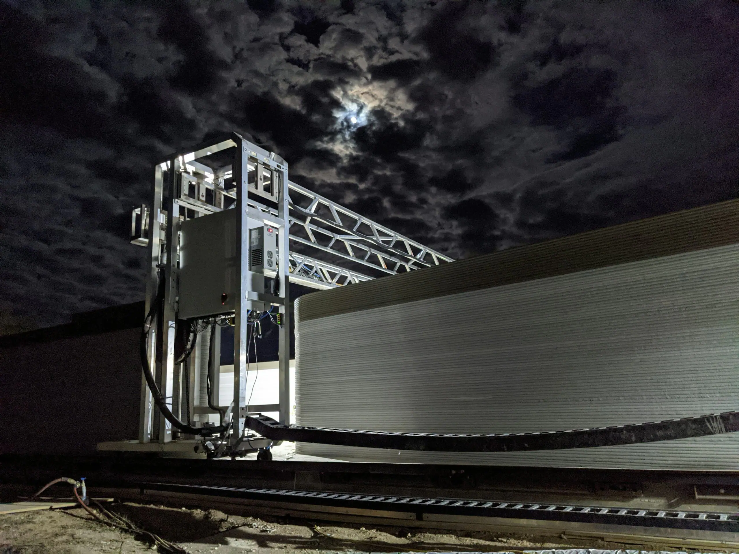 Robotic gantry 3D printer forming a ribbed concrete wall at night under moonlit clouds