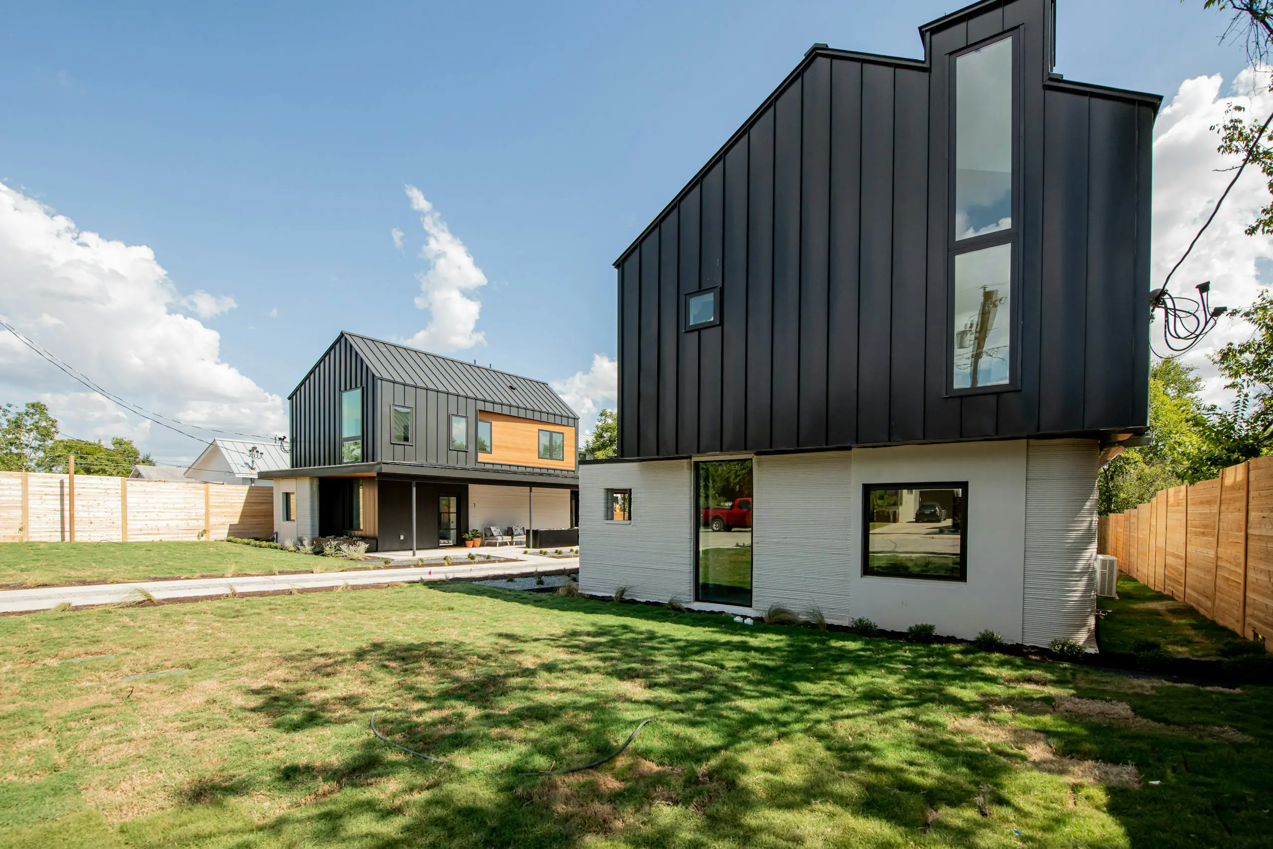 Pair of modern design‑build homes with black standing‑seam metal cladding, wood accent panel, and landscaped front yard