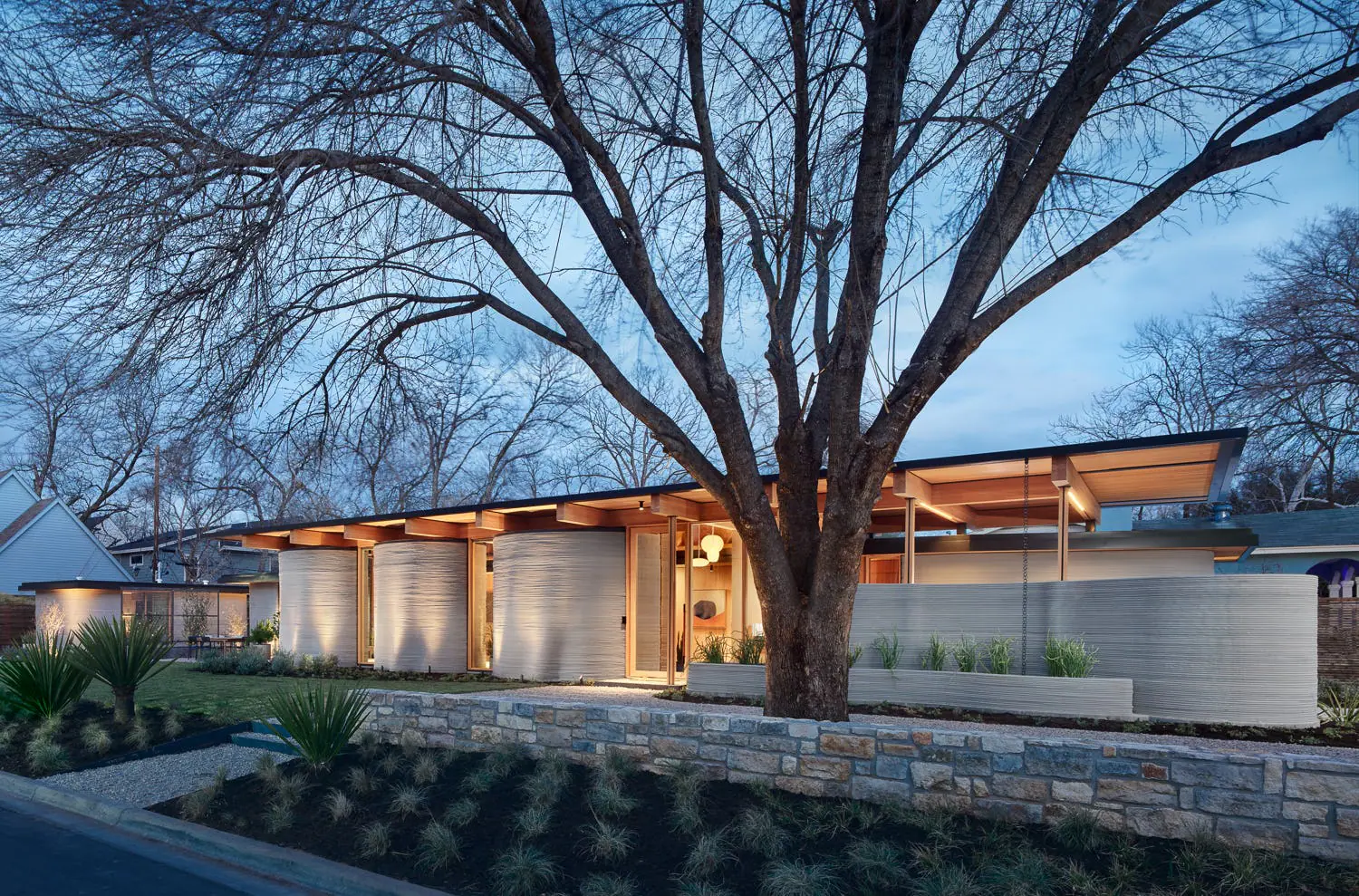 Evening view of a modern 3D-printed home with curved concrete walls, exposed wood roof and landscaped front yard