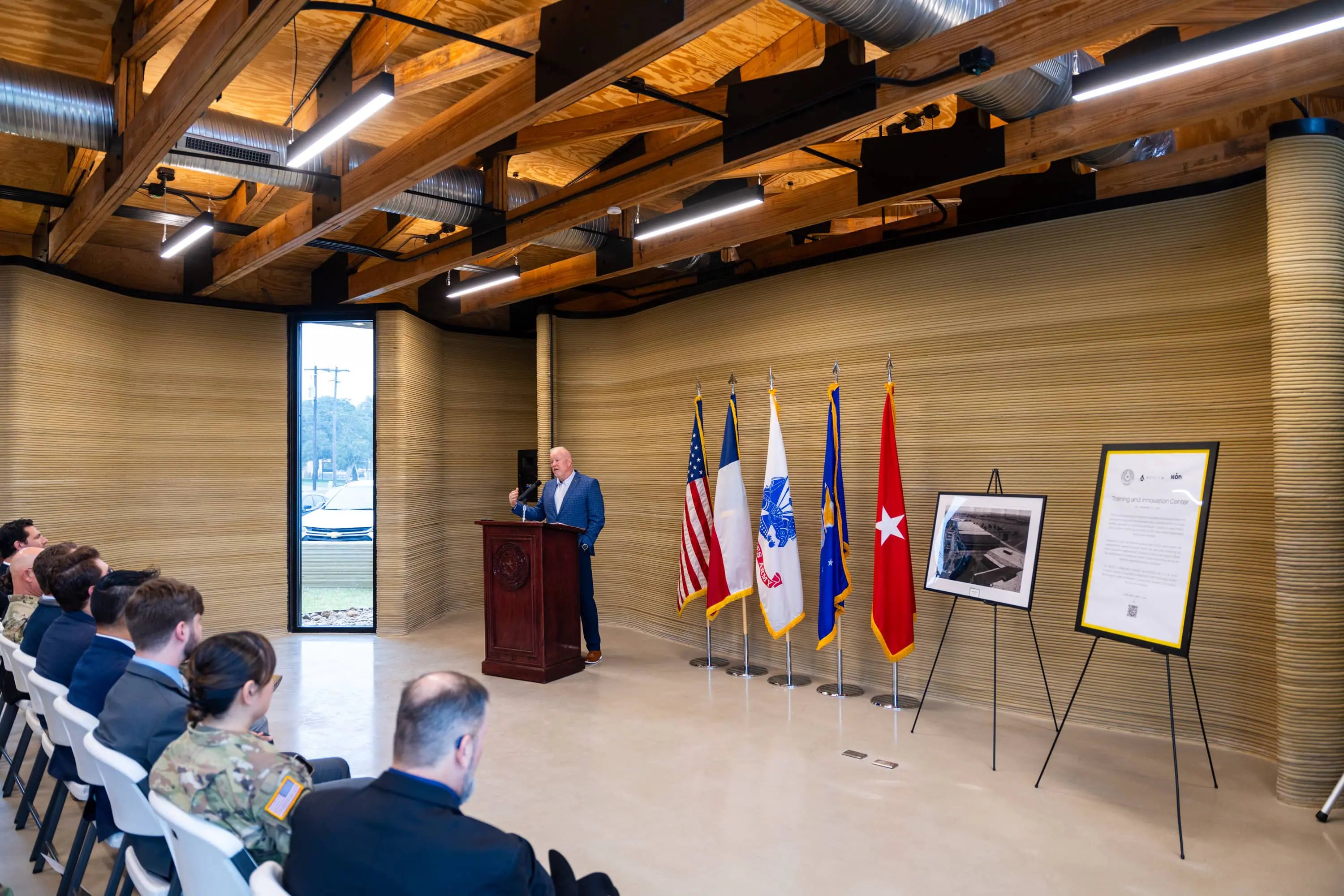 Speaker at podium in a building lobby with 3D-printed ribbed concrete walls, exposed timber ceiling and audience