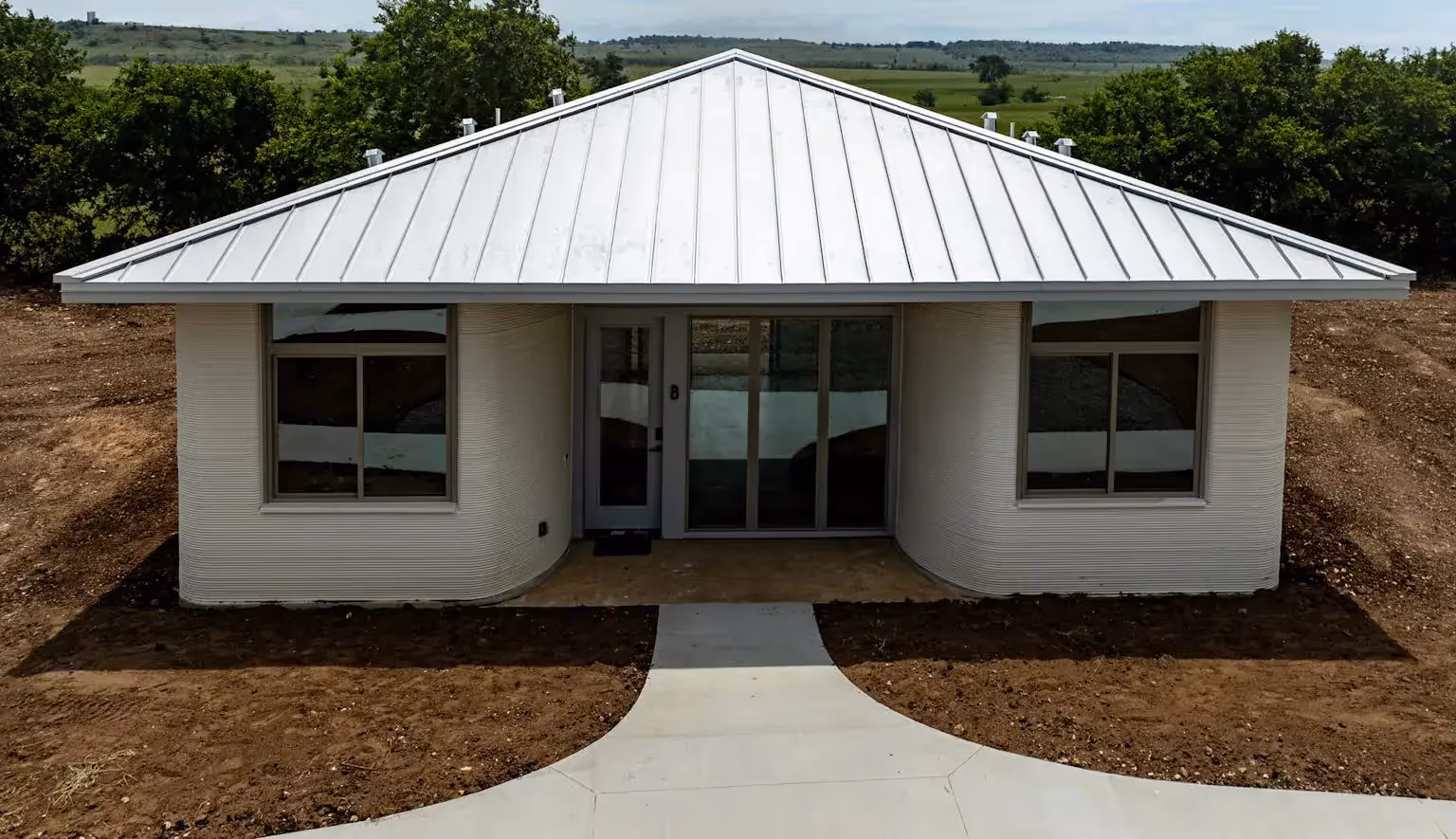 3D-printed single-story home with ribbed curved walls, metal roof and sliding-glass entry on a cleared construction site