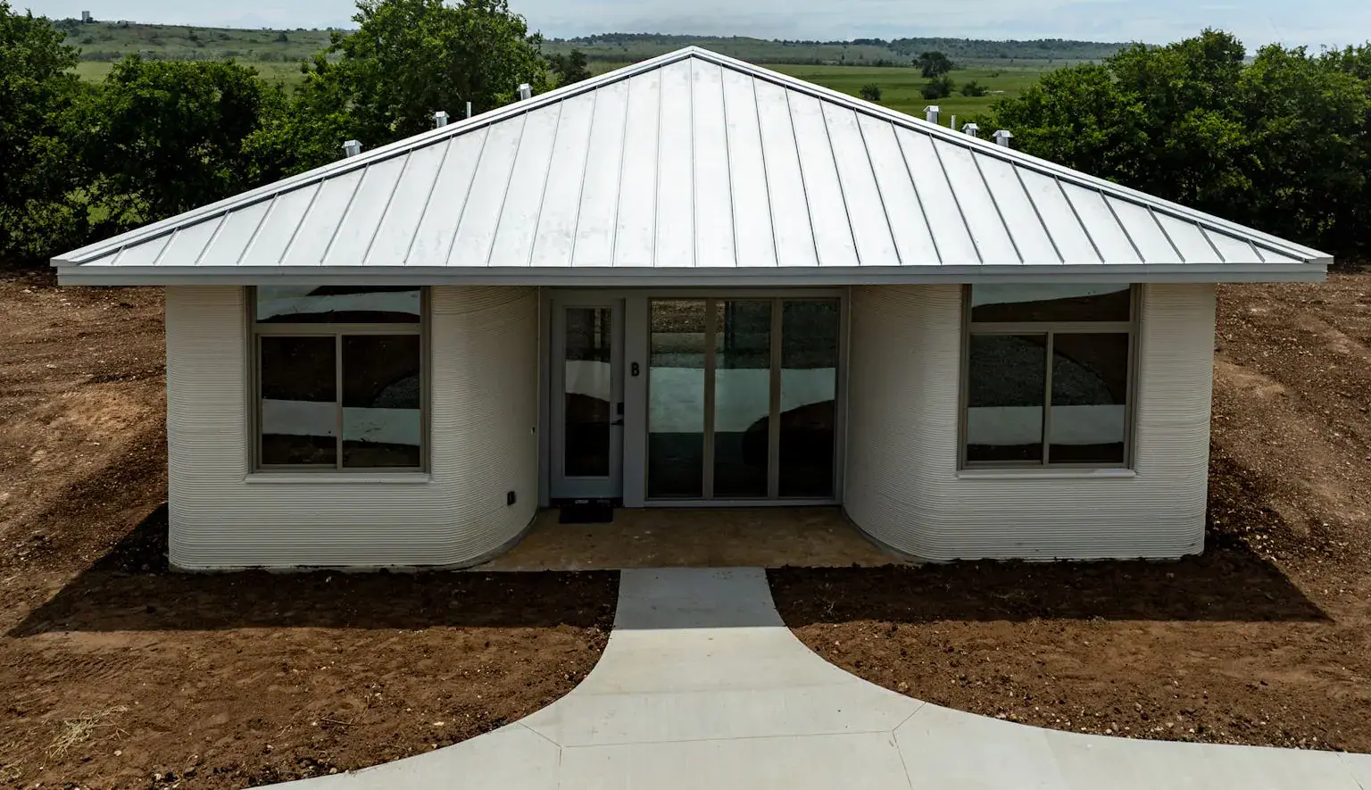 3D-printed single-story home with ribbed curved walls, metal roof and sliding-glass entry on a cleared construction site