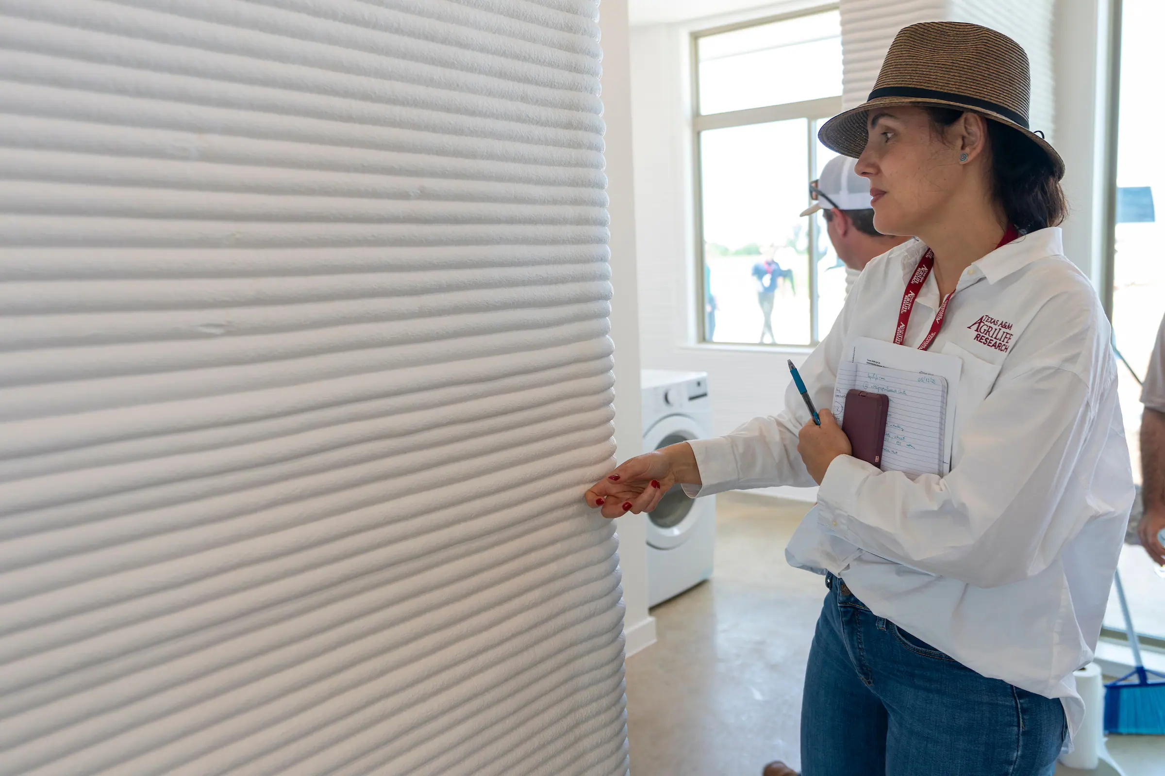 Woman with hat and notebook inspecting ribbed 3D-printed concrete wall