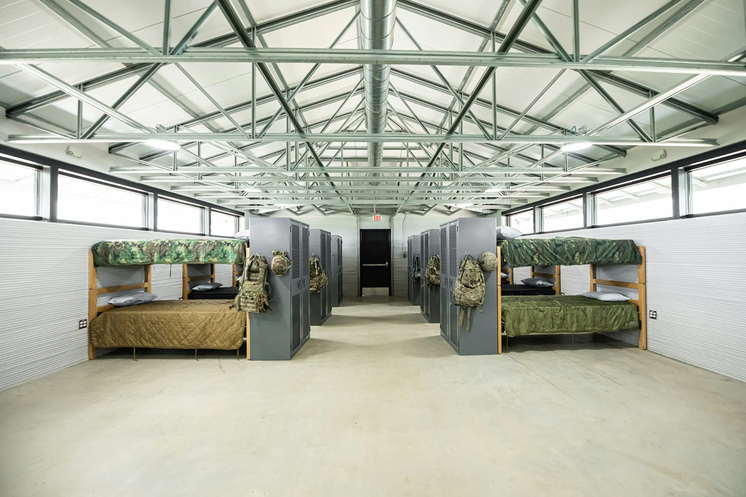 Communal bunkroom with lockers, camouflage bedding and exposed steel trusses illustrating modular housing