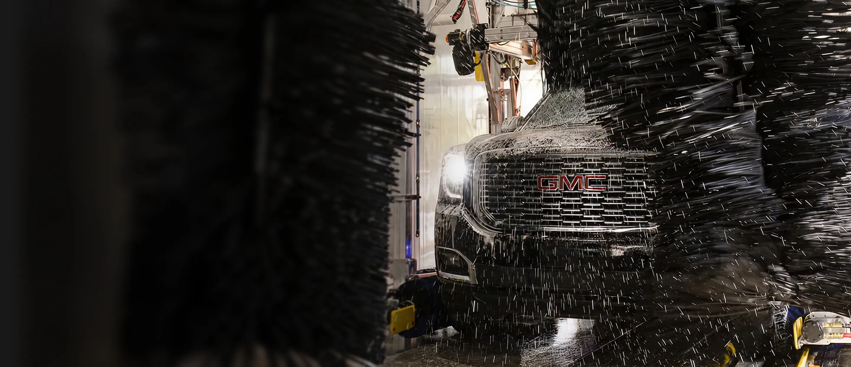 Front of a black GMC vehicle being washed inside an automatic car wash with water and brushes.