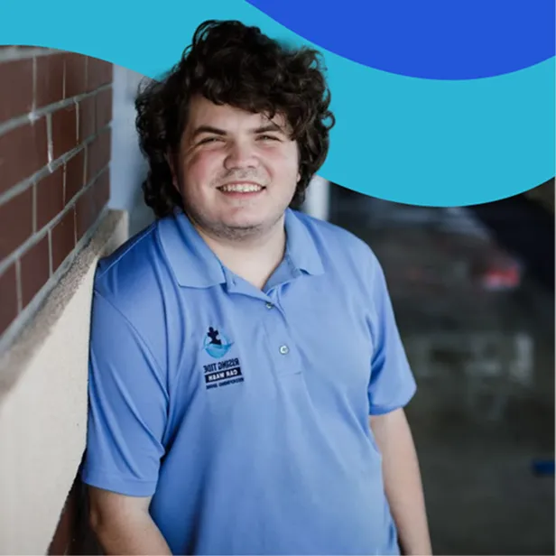 Smiling young man with curly hair leaning against a brick wall, wearing a light blue polo shirt with a logo.