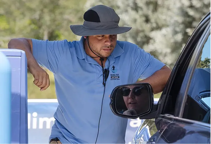 A man wearing a wide-brimmed hat and a blue polo shirt leans on a car window while talking to the driver reflected in the side mirror.