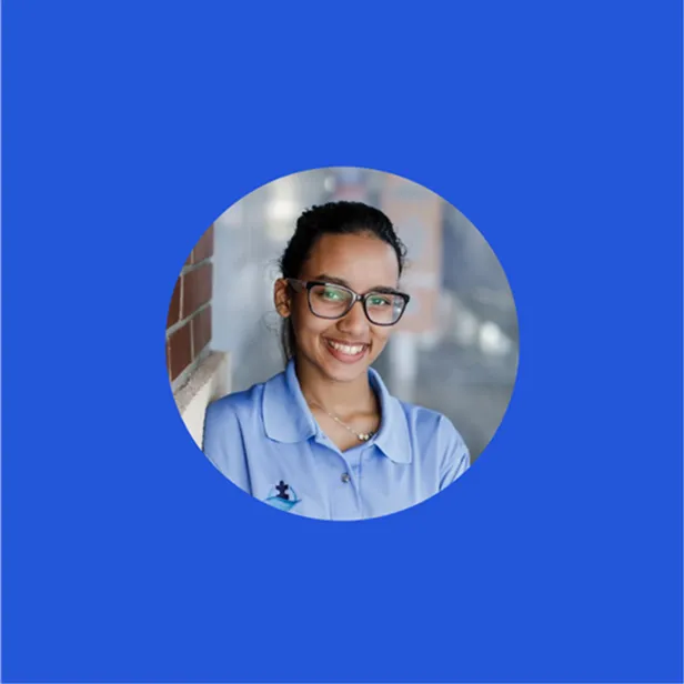 Smiling woman with glasses wearing a light blue polo shirt leaning against a brick wall.