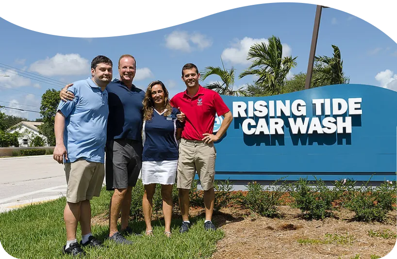 Four people standing together and smiling in front of a large blue sign that reads 'Rising Tide Car Wash' on a sunny day.