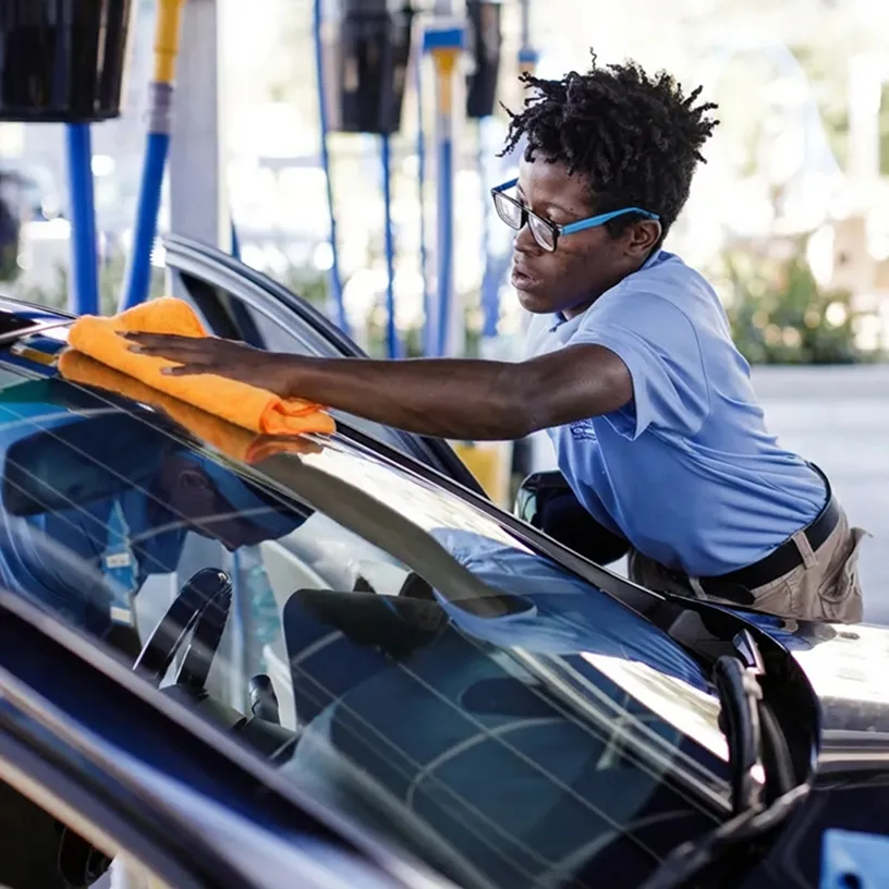Person in blue shirt and glasses cleaning a car windshield with an orange cloth.