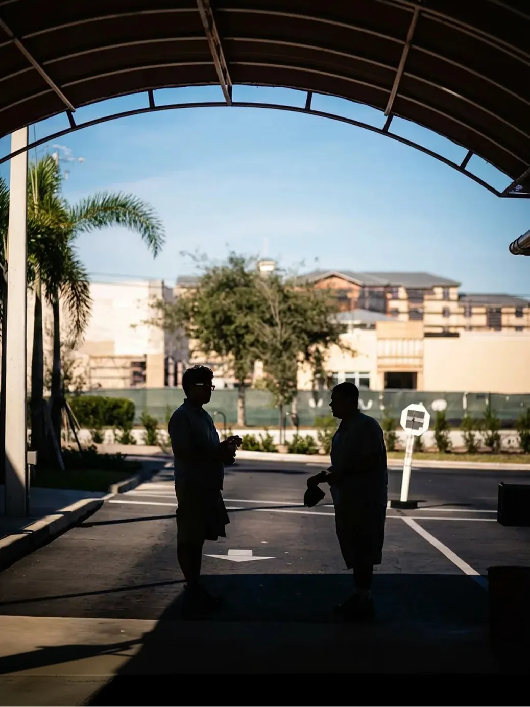 Silhouettes of two people standing under a large canopy with buildings, trees, and a clear sky in the background.