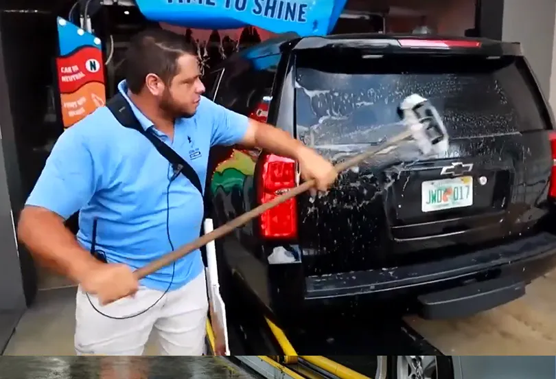 Man in blue shirt washing the back of a black Chevrolet SUV with a long-handled brush at a car wash.