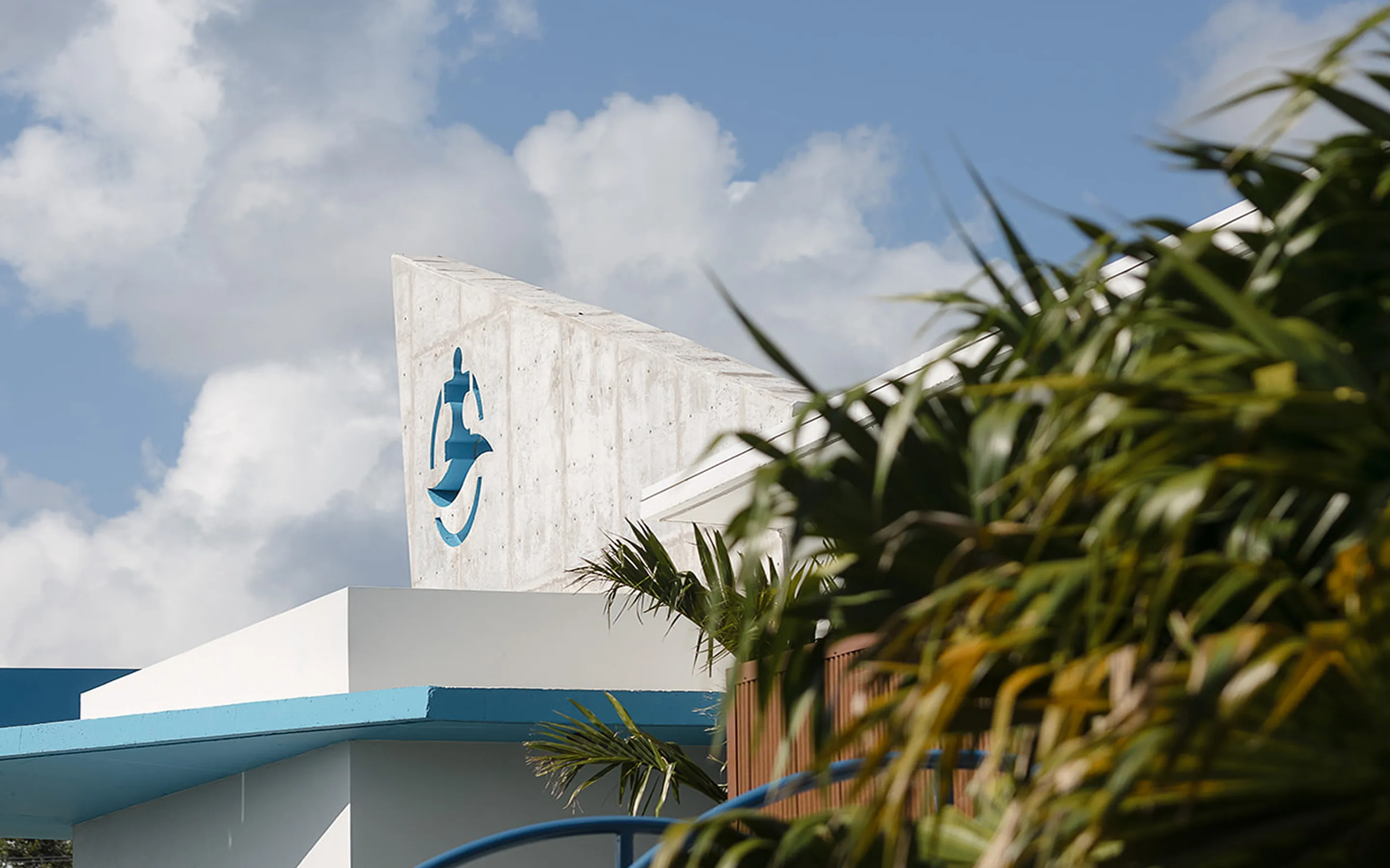 Modern building corner with concrete wall featuring a blue abstract figure logo and tropical plants in the foreground under a partly cloudy sky.