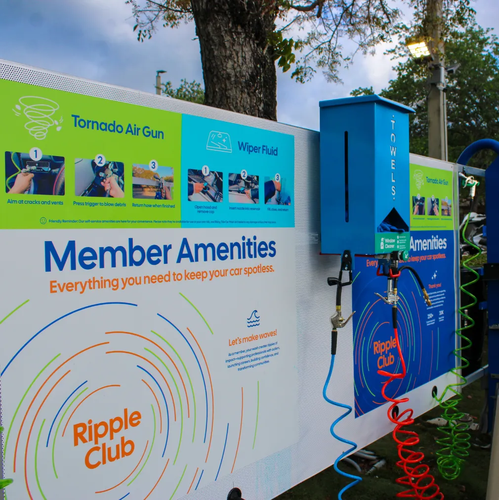 Self-service car cleaning station with instructions for using tornado air gun and wiper fluid along with a blue towel dispenser, colorful coiled hoses, and a sign for Ripple Club member amenities.