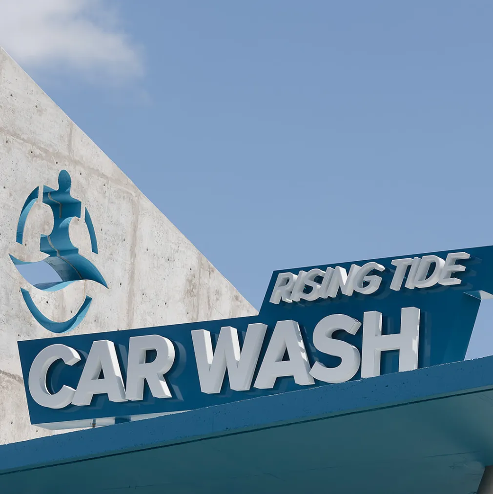 Sign reading 'Rising Tide Car Wash' against a blue sky with a stylized person and waves logo on a concrete wall.